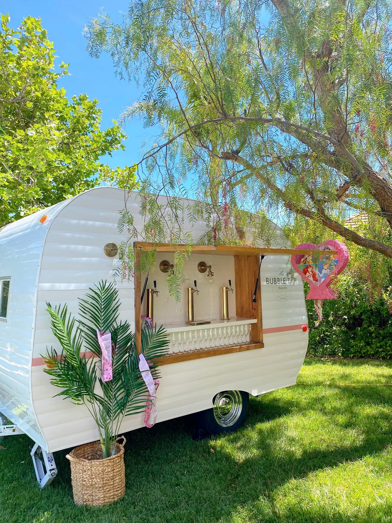 A white mobile bar trailer called "The Bubble Tea Trailer" serving drinks, set outdoors under a tree with greenery and a blue sky. There is a pink heart-shaped decoration, a potted plant, and glasses on the counter.