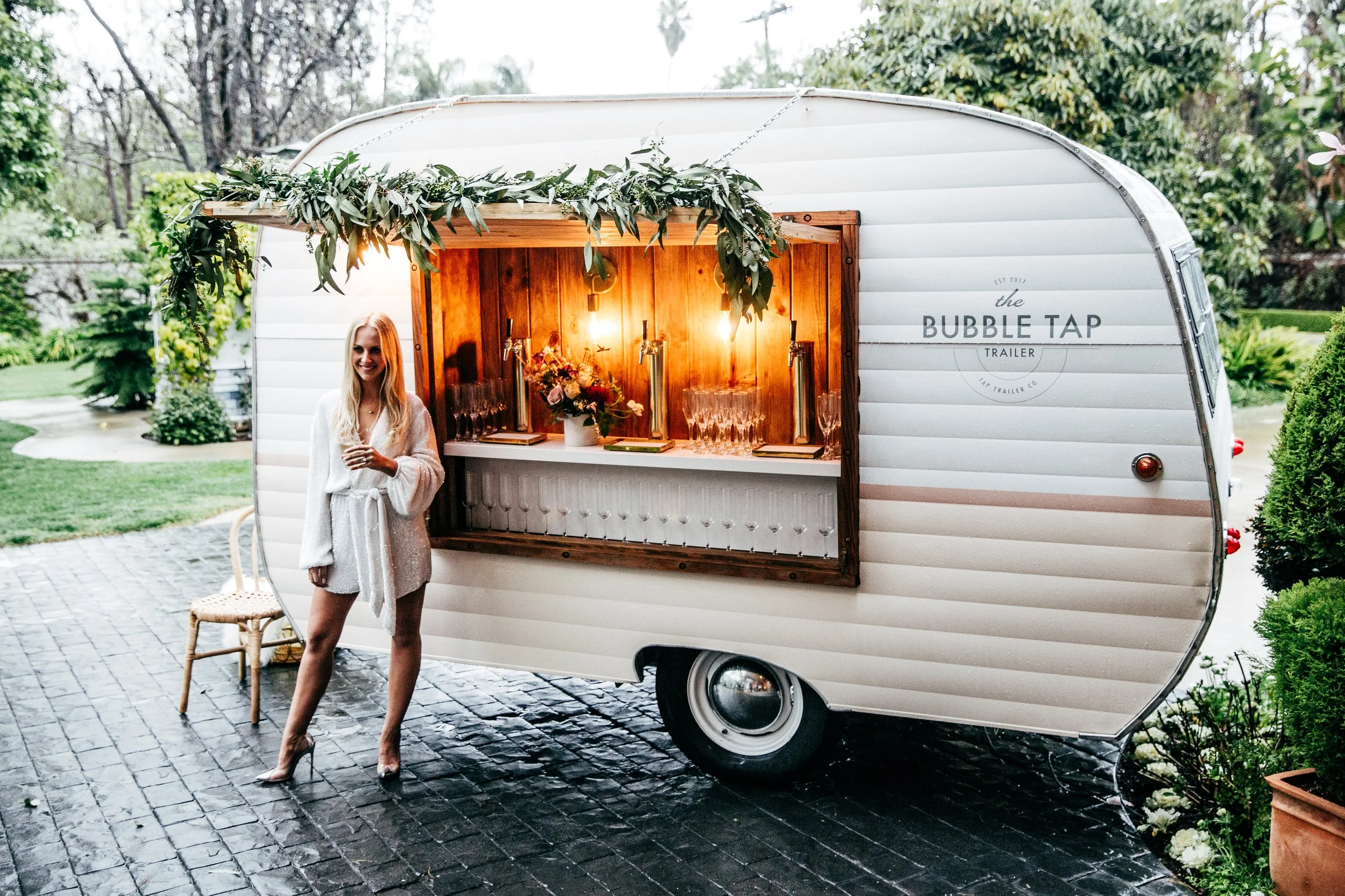A woman standing next to a mobile bar trailer named 'the BUBBLE TAP' with a wooden and white exterior, decorated with greenery and flowers, serving drinks on a gravel path in a lush outdoor setting.
