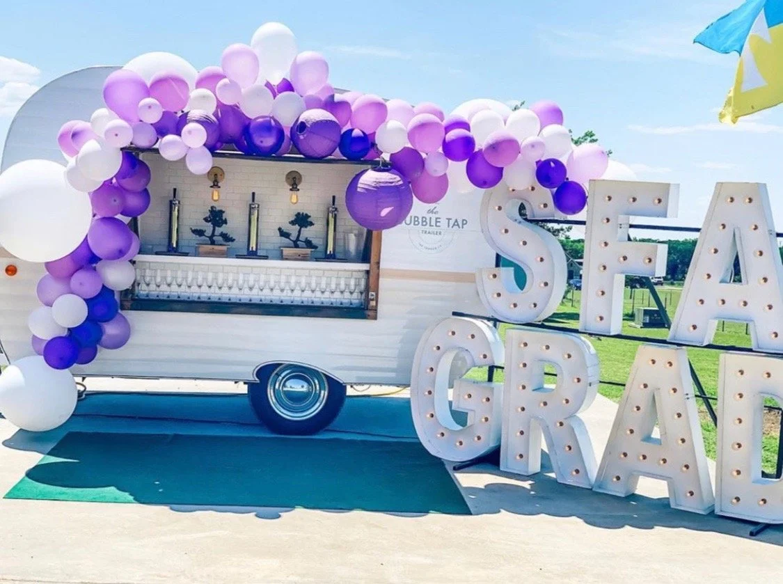 Decorated ice cream trailer with purple and white balloons, large illuminated letters spelling 'SEA GRASS,' and a green mat on the pavement.