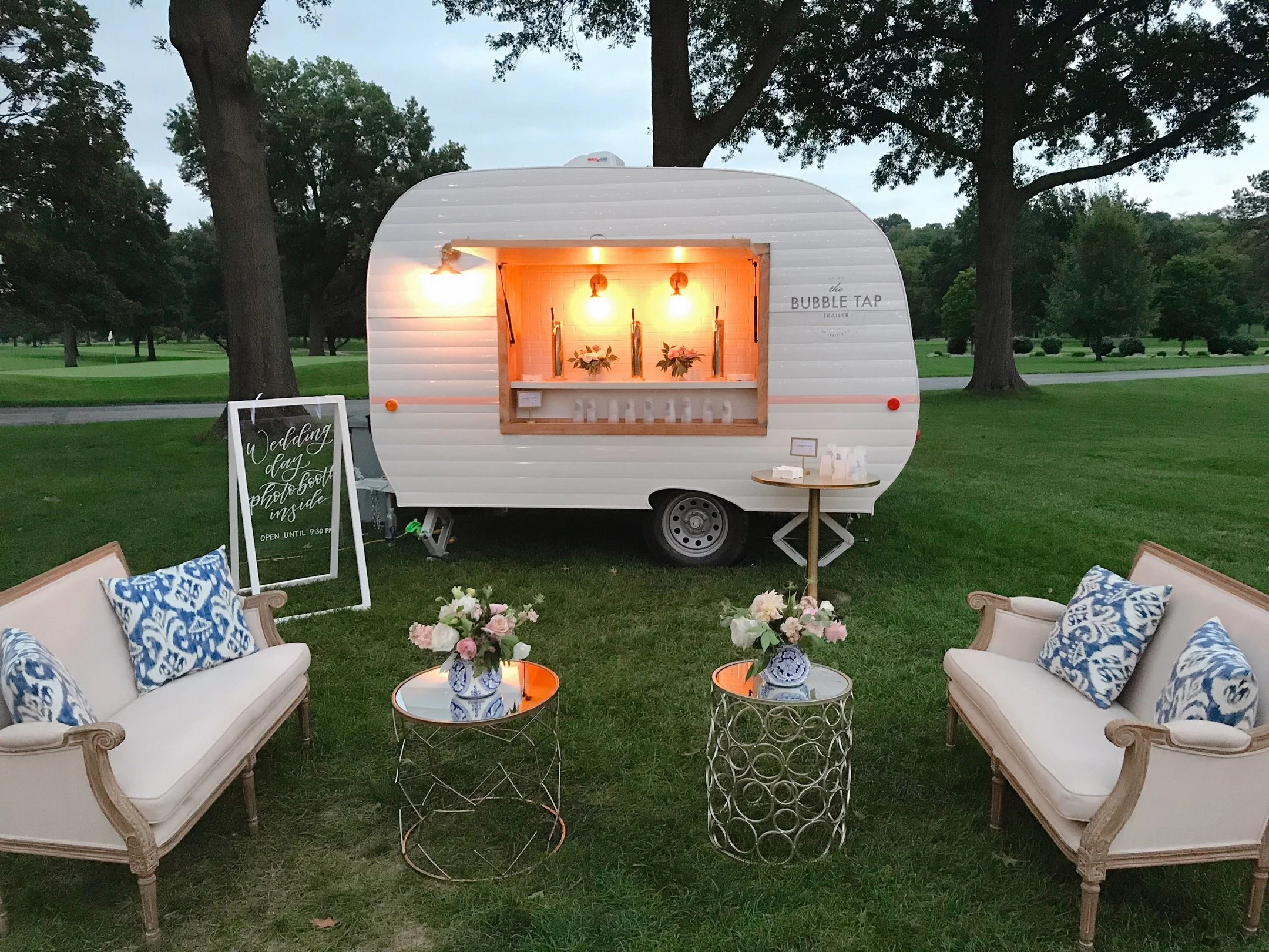 A white mobile bar trailer named 'Bubble Tap' set up outdoors in a grassy park with trees in the background. Two vintage-style sofas with blue and white pillows are arranged in front of the trailer, with small round glass-topped tables holding flower arrangements between them. A chalkboard sign on a stand indicates a wedding day photo booth inside. The trailer has a window with bottles and flowers inside, lit with warm white lights.