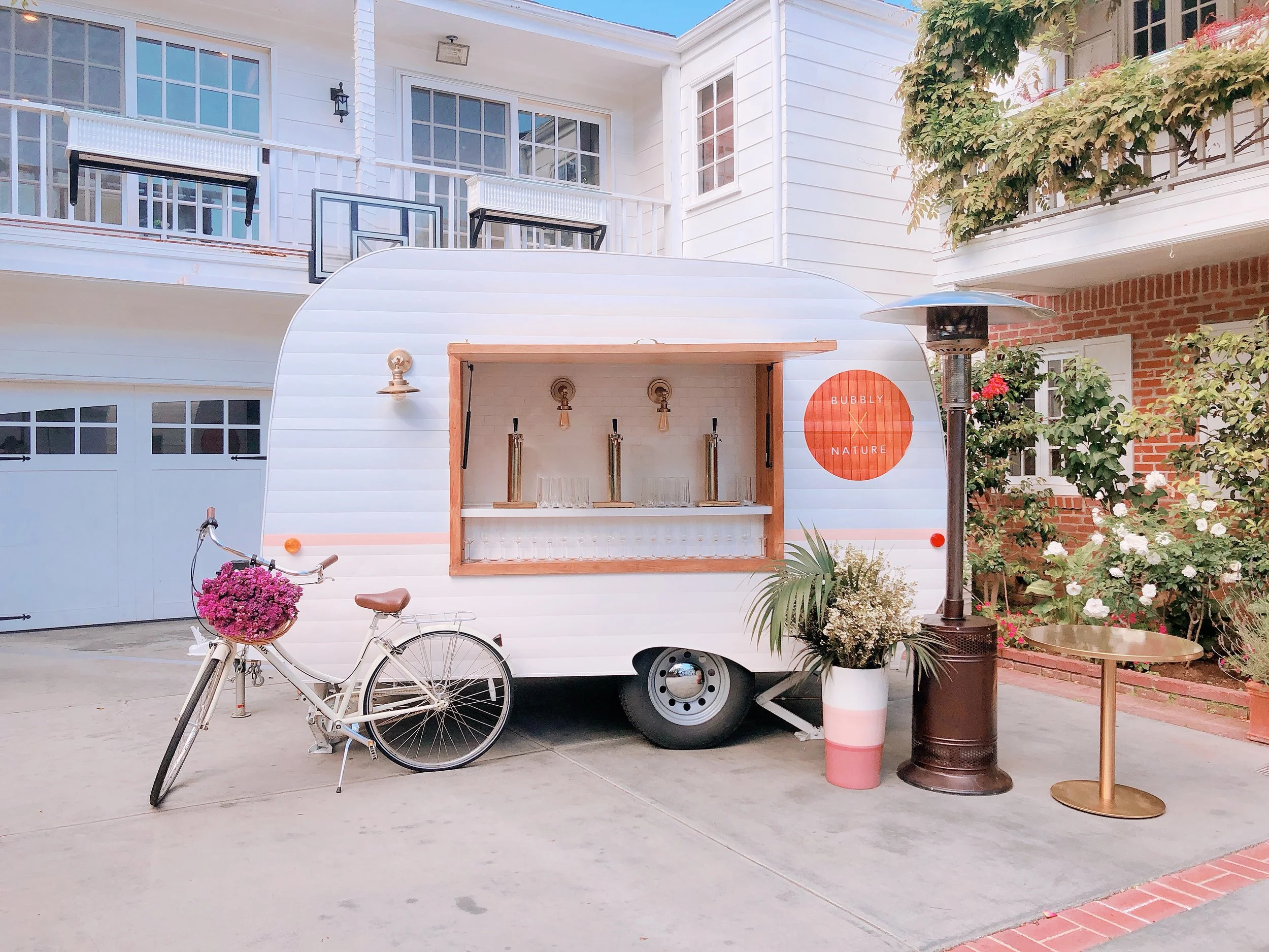 A white mobile coffee or drink stand with a small window, set up on a driveway near a brick house. There is a bicycle with a pink flower basket parked in front. The stand has three taps and glassware on a shelf. Nearby are a tall heater, a potted plant, and a small round table with a gold-colored top. The background features greenery and a house with balconies and basketball hoops.