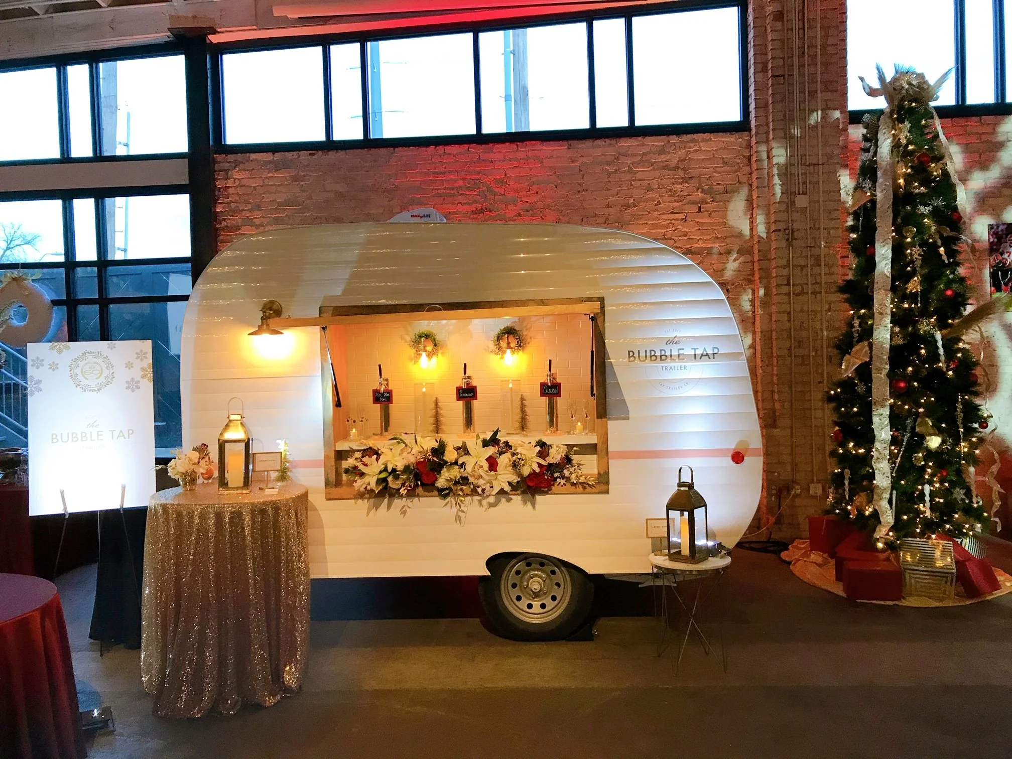 Festive holiday scene featuring a white vintage trailer decorated for Christmas, with a sign reading 'the Bubble Tap'. The trailer has three beer taps, floral decorations, and warm lighting. A decorated Christmas tree with gold and red ornaments and ribbons stands beside it, with wrapped presents underneath. To the left, a round table with a glittery tablecloth and a lantern adds to the festive atmosphere, in an industrial-style space with brick walls and large windows.