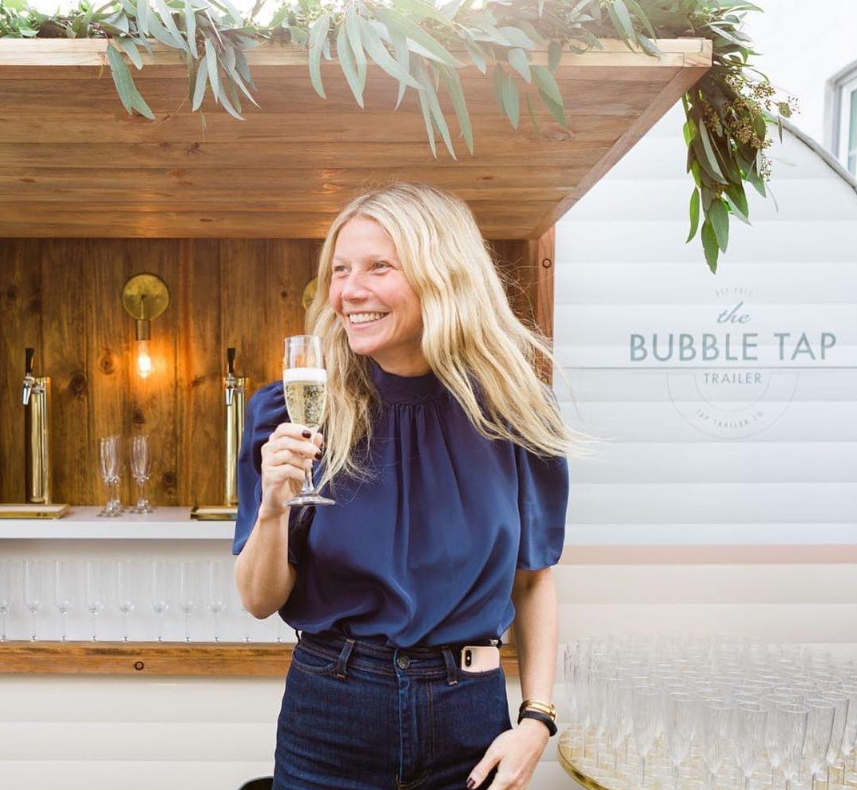 A smiling woman with blonde hair wearing a navy blue blouse and dark jeans, holding a glass of champagne at a bar or tasting event.