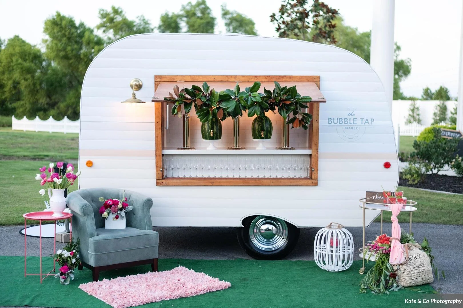 A mobile bar trailer called 'the Bubble Tap' decorated with greenery and flowers, set up outdoors with a seating area including a plush chair, a small pink table with flowers, and a side table with drinks, on a green rug, surrounded by trees and plants.