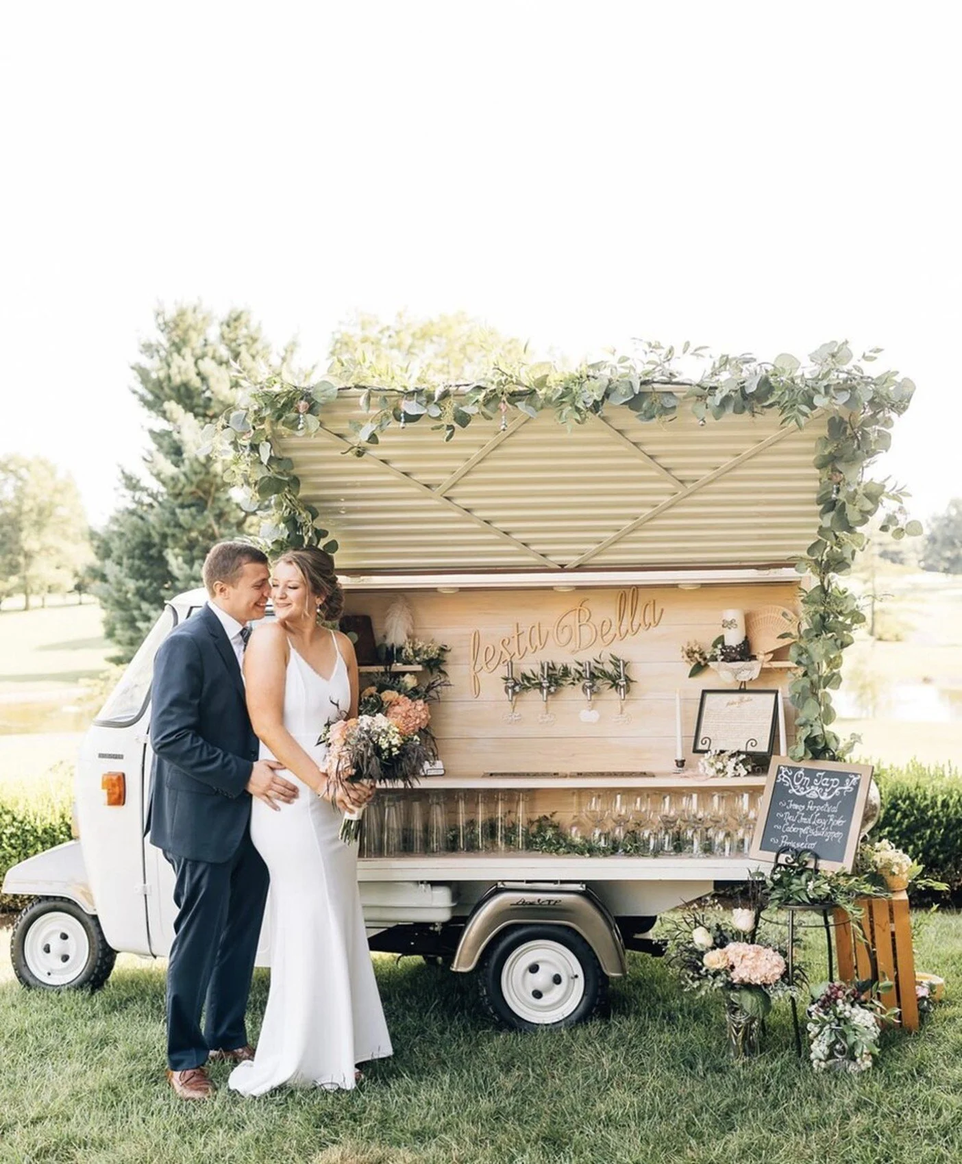 A newlywed couple in wedding attire standing outside near a decorated mobile bar with a sign that says "Festa Bella" on a sunny day with greenery and trees in the background.
