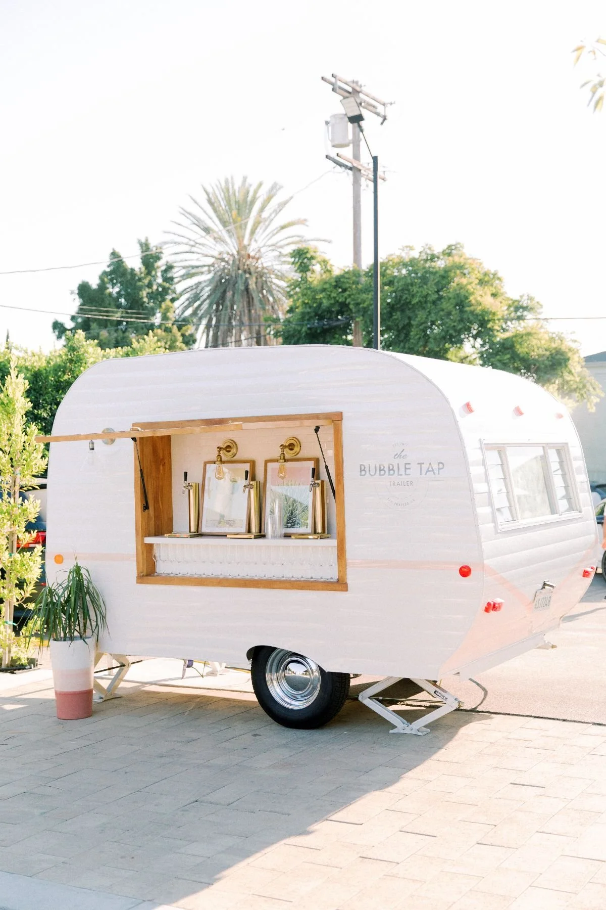Pink and white mobile bubble tea trailer with a window serving area, decorated with framed pictures, located on a sidewalk with green plants and trees in the background.