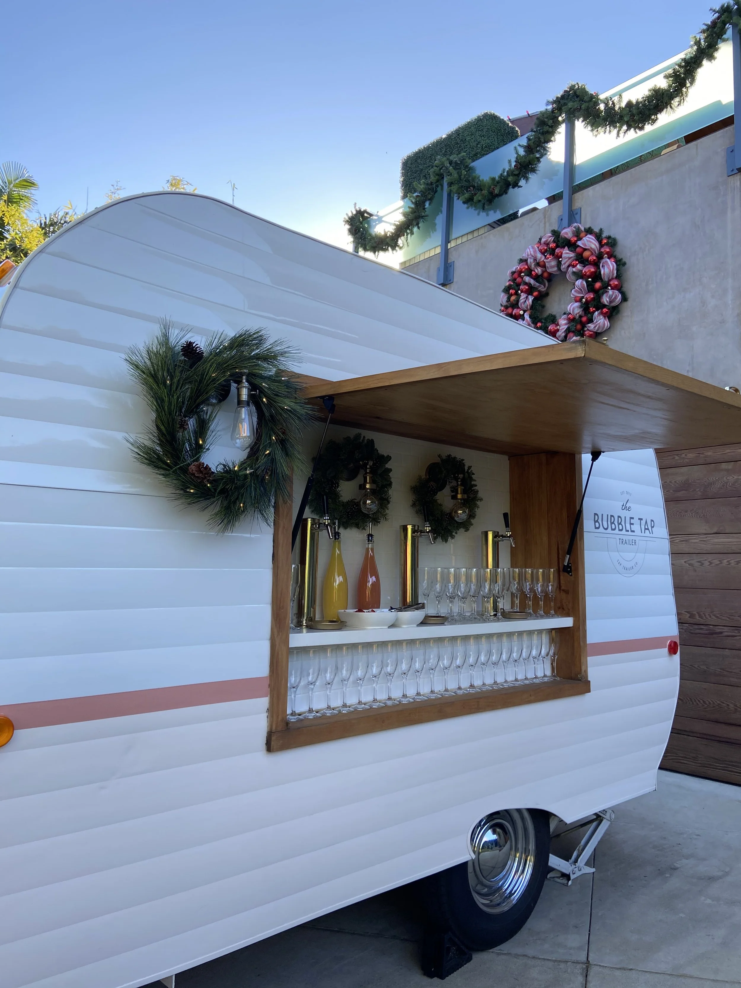 A white vintage-style trailer decorated for Christmas with wreaths, ornaments, and garlands. It has a wooden serving window with drink dispensers, glasses, and holiday decorations.