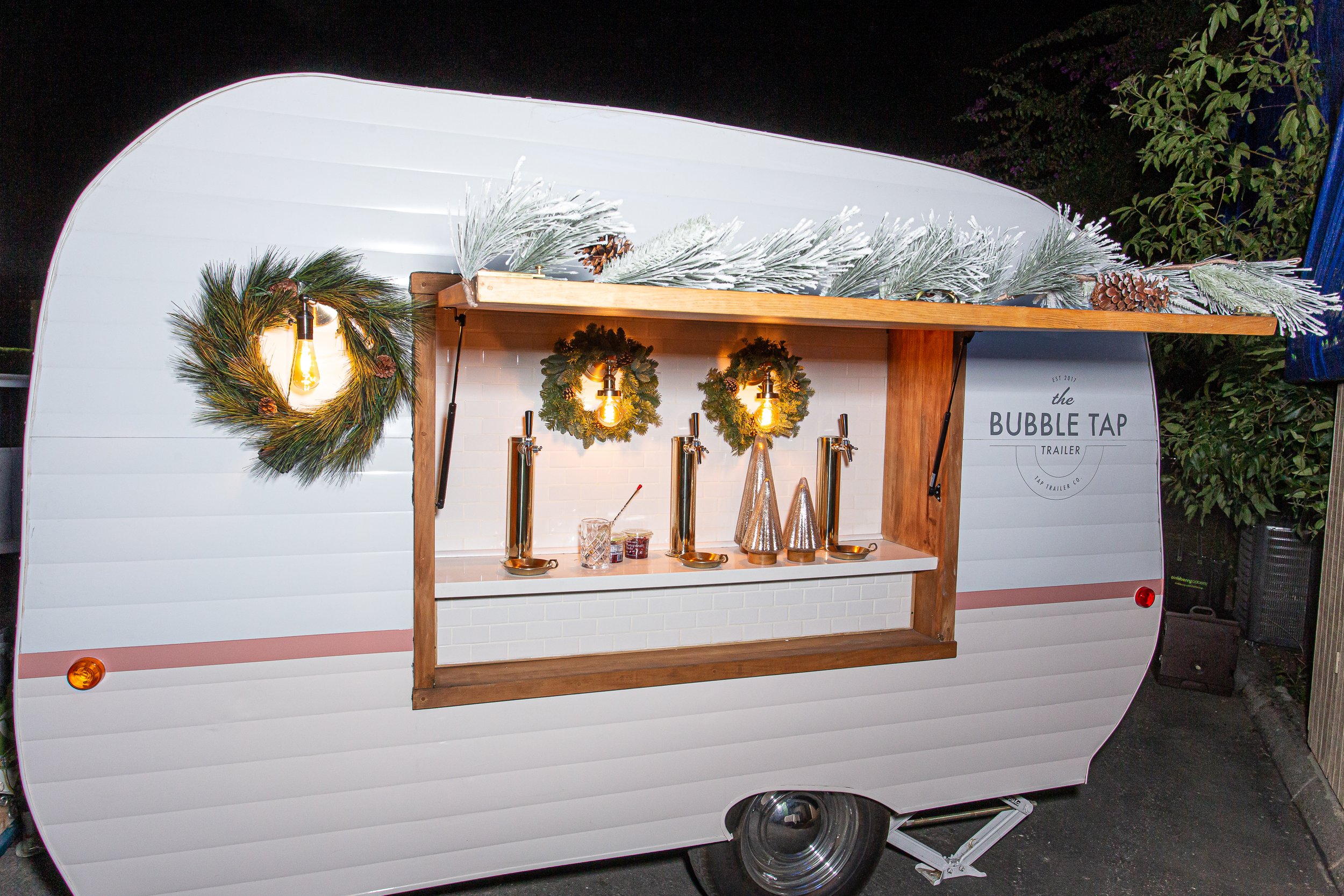 A white mobile bar with a wooden front panel, decorated with holiday greenery and wreaths, featuring four beer taps, jars of jam, and hanging light bulbs inside.