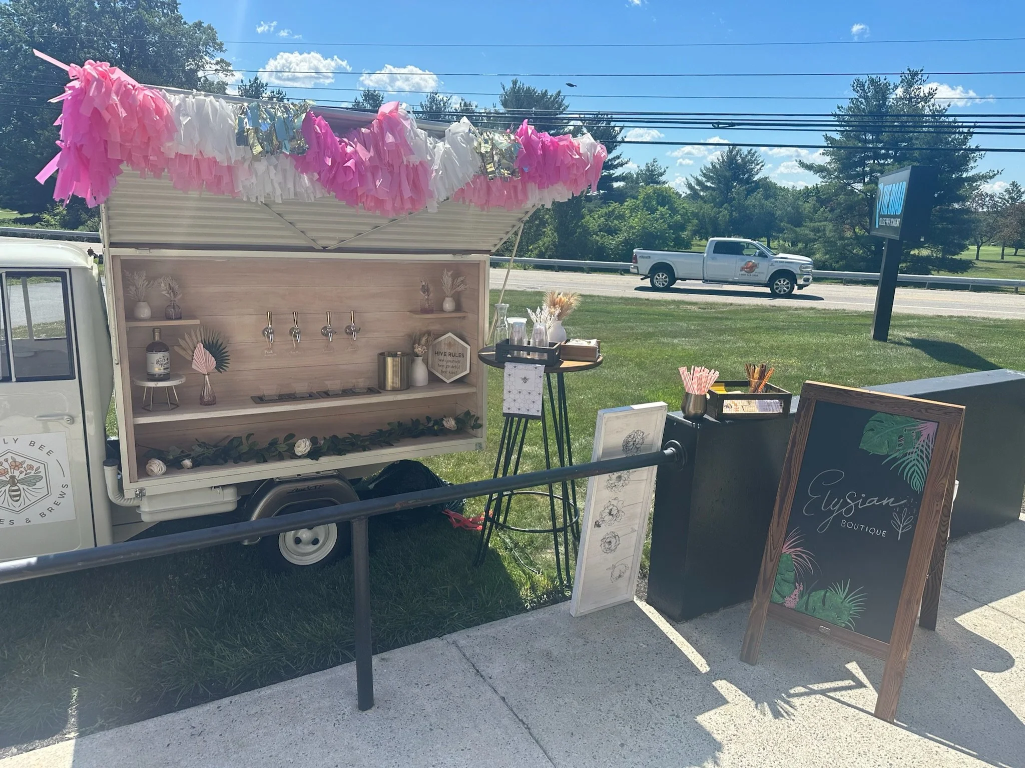 A mobile bar setup with a pink and white fringe canopy, decorated with flowers and greenery, set up outside on a grassy area. There are signs, a small blackboard sign reading 'Elysian Boutique,' and various drinkware and decorative items displayed.