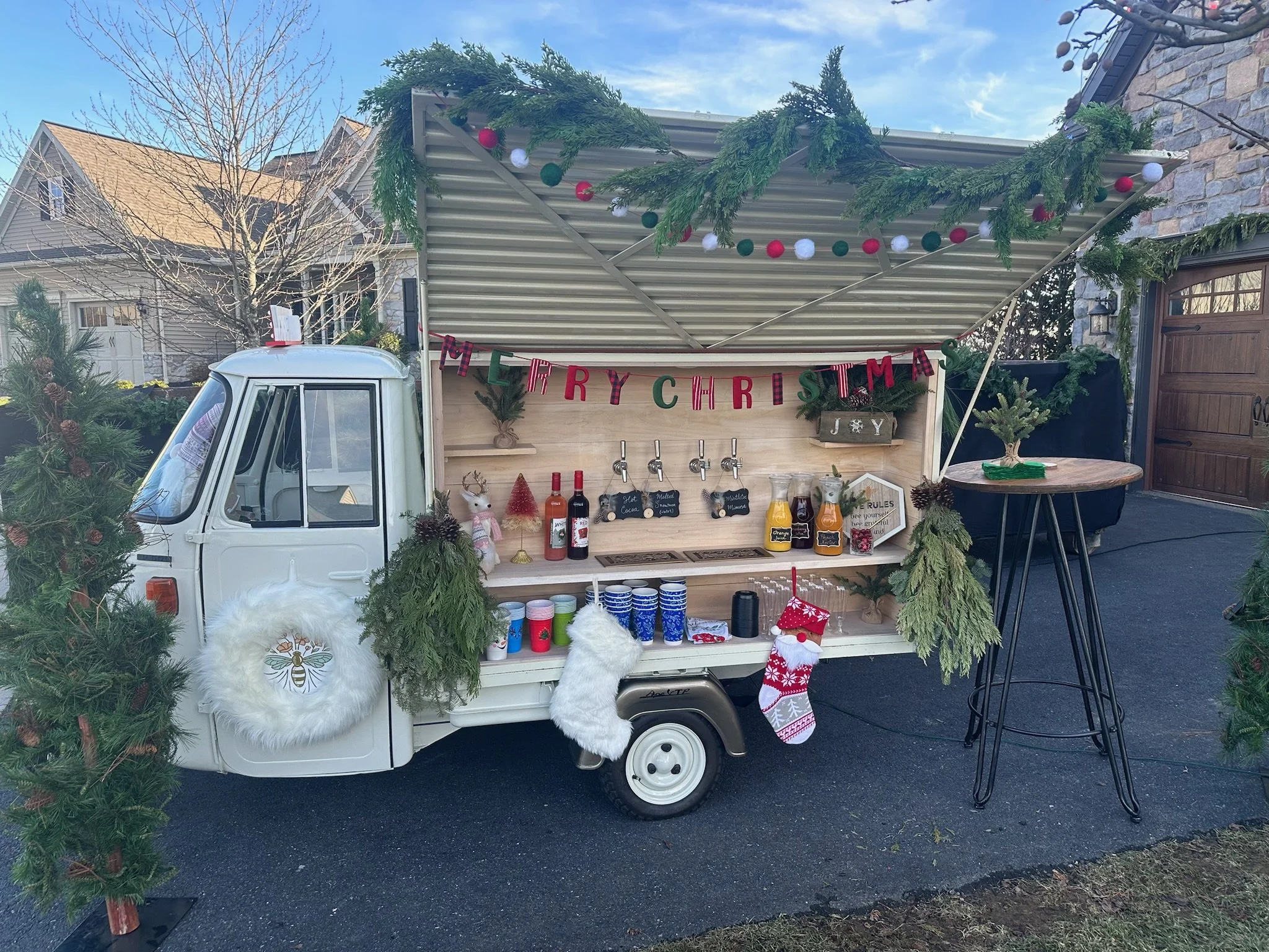 A decorated mobile Christmas stand on wheels with holiday ornaments, a garland, and a "MERRY CHRISTMAS" banner, set in a residential area with houses and bare trees in the background.