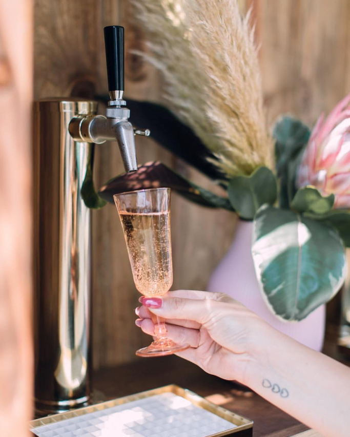 A hand with pink nails holding a pink champagne glass under a tap dispensing sparkling rose wine or champagne, with a blurred floral arrangement in the background.