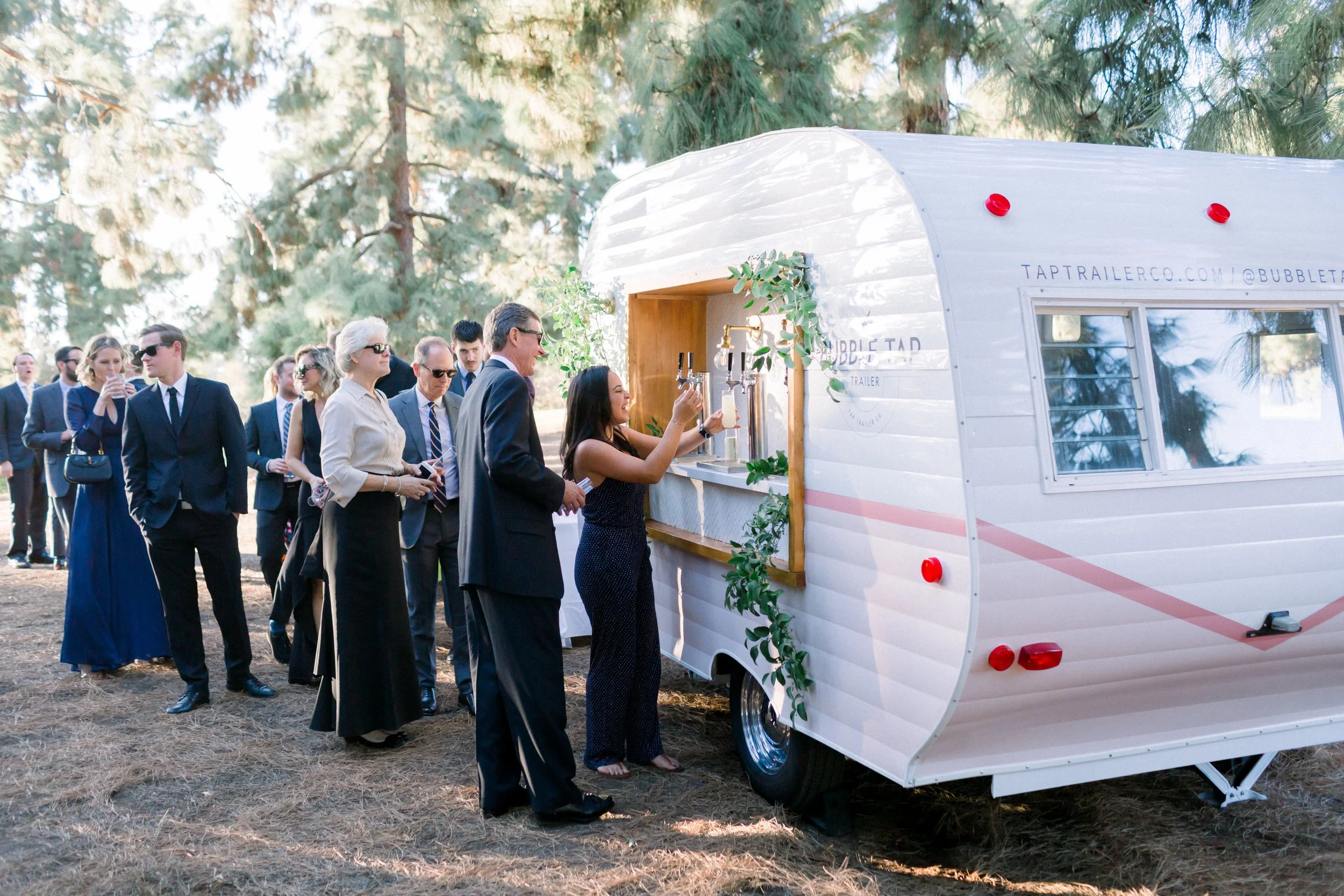 People in formal attire standing in line at an outdoor beverage trailer with a woman pouring drinks.