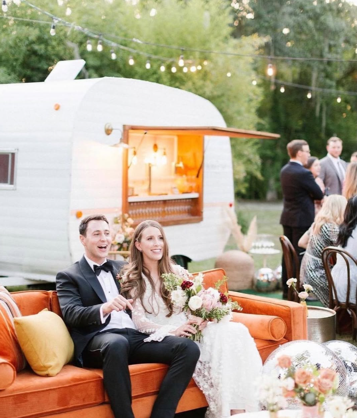 A wedding reception outdoors with a couple sitting on an orange vintage sofa, the bride holding a bouquet of flowers, and guests in the background near a white trailer with a serving window, string lights overhead, and decorative items on the ground.