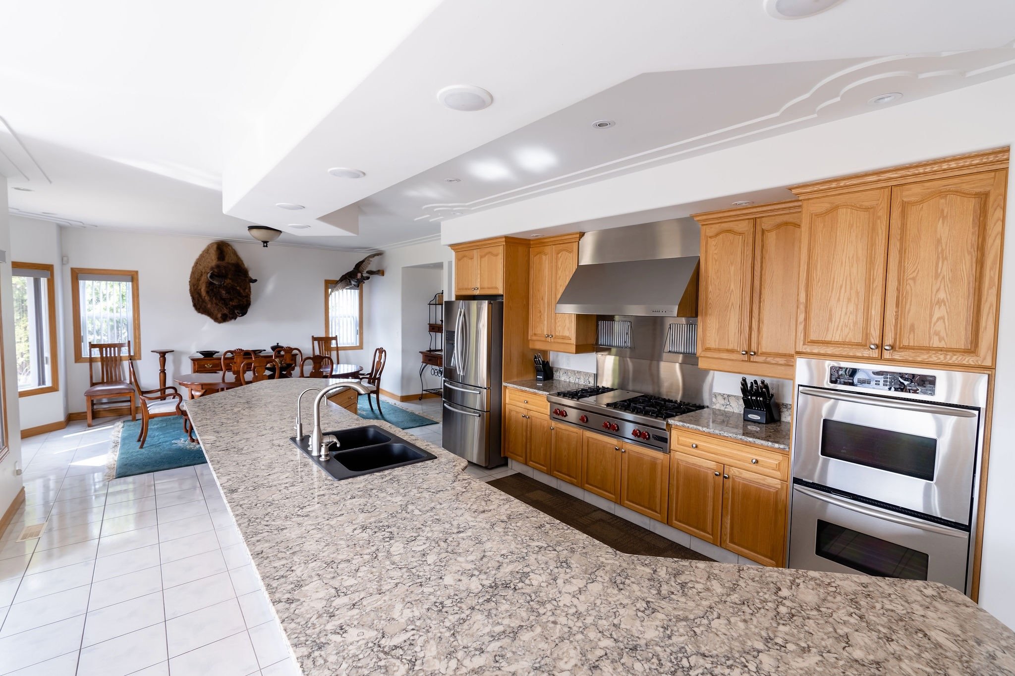 Modern kitchen with granite countertops, wooden cabinets, stainless steel appliances including a double oven and refrigerator, and a breakfast bar. In the background, there is a dining area with a table, chairs, and framed windows.