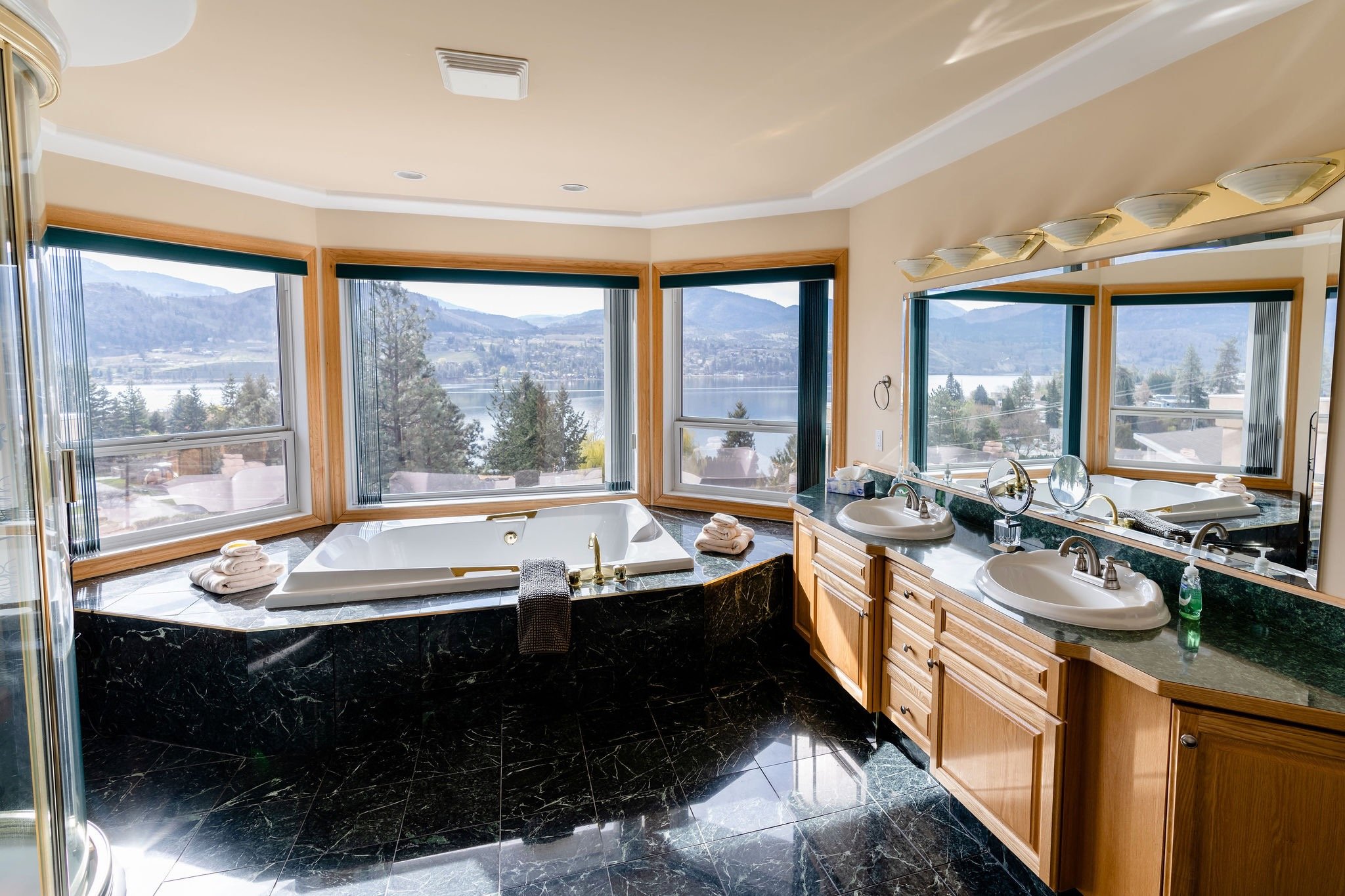 Bathroom with a large corner bathtub near three windows with scenic mountain and lake views, a double sink vanity, and a large mirror.