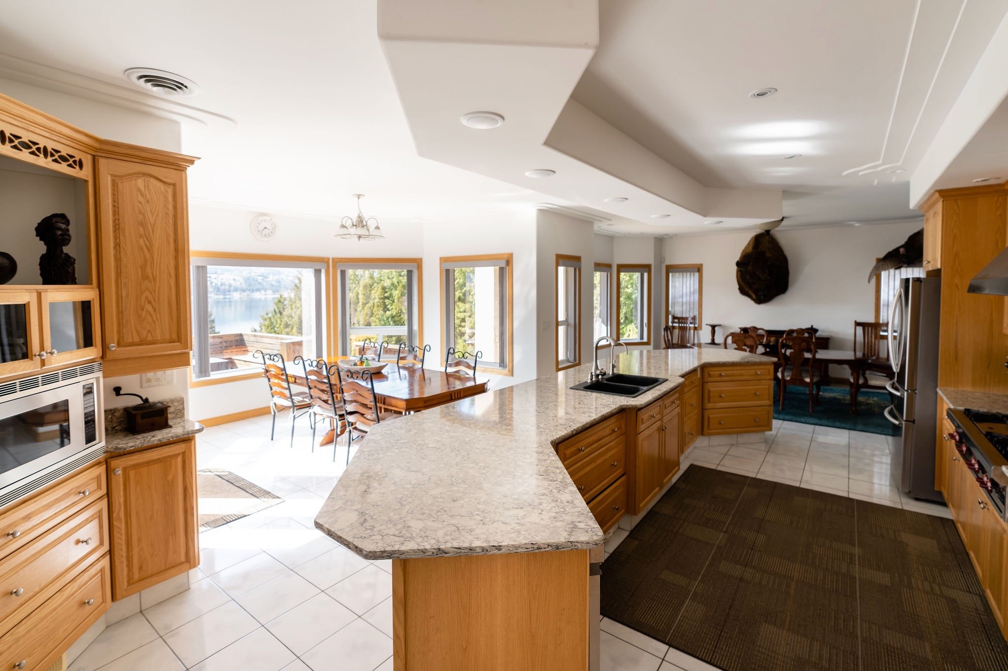 Open-concept kitchen with wooden cabinets, granite countertops, and a large island with a sink, leading into a dining area with a wooden table and chairs near large windows showing a view of a lake and trees.