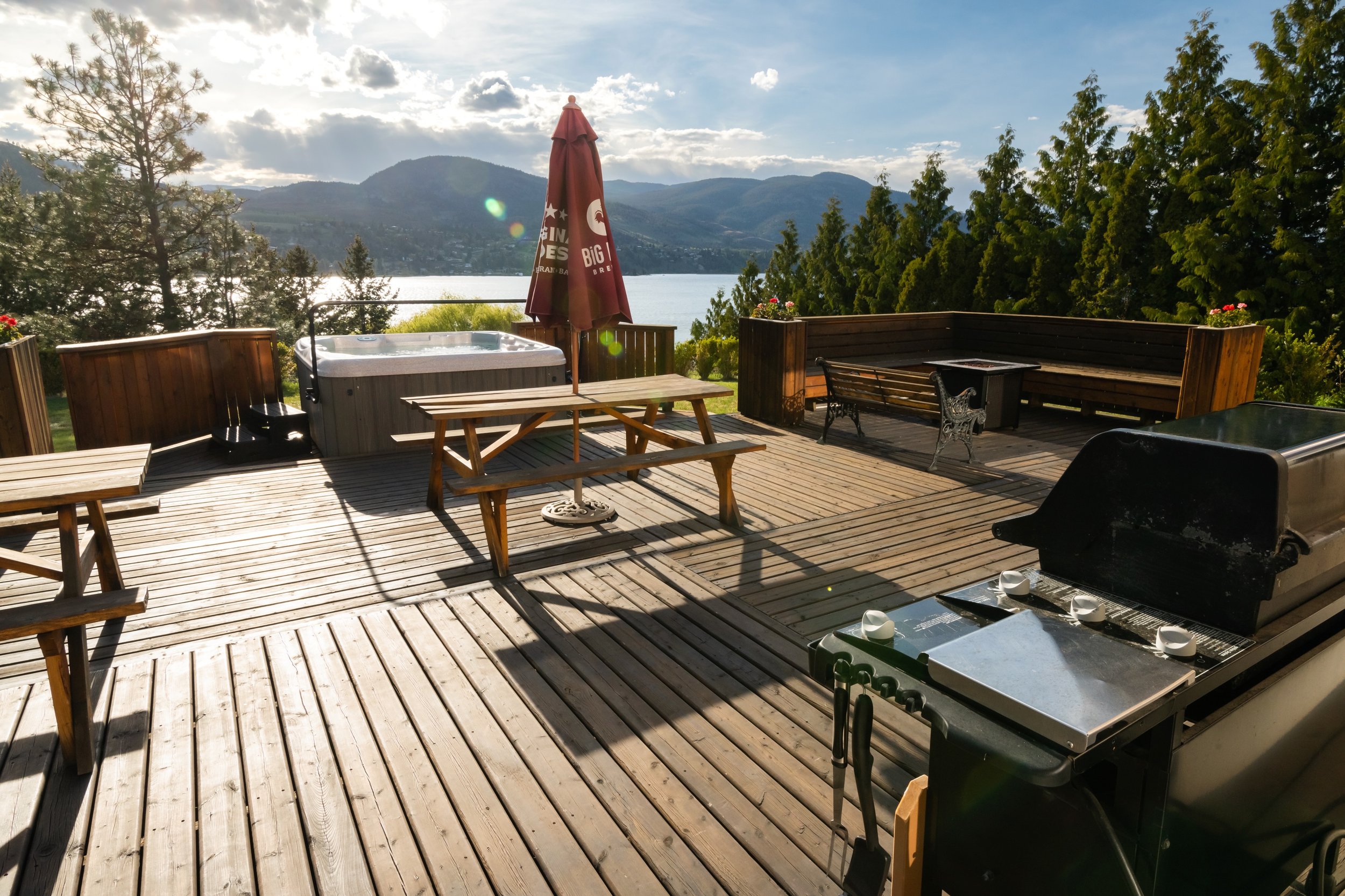 A spacious wooden deck overlooking Skaha Lake with mountains in the background. The deck has a hot tub, a picnic table with an umbrella, and some outdoor furniture, surrounded by lush green trees.