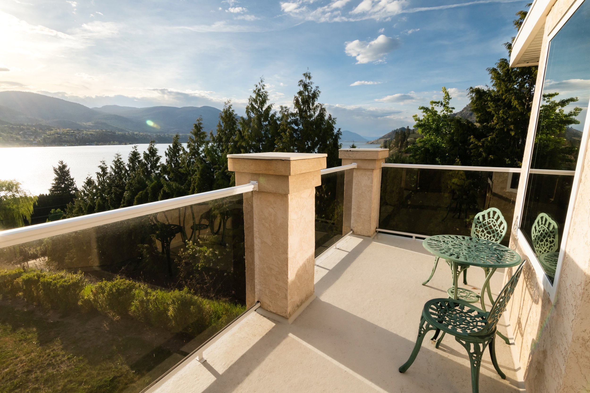 Balcony with green metal table and chair overlooking Skaha Lake and mountains under a partly cloudy sky.