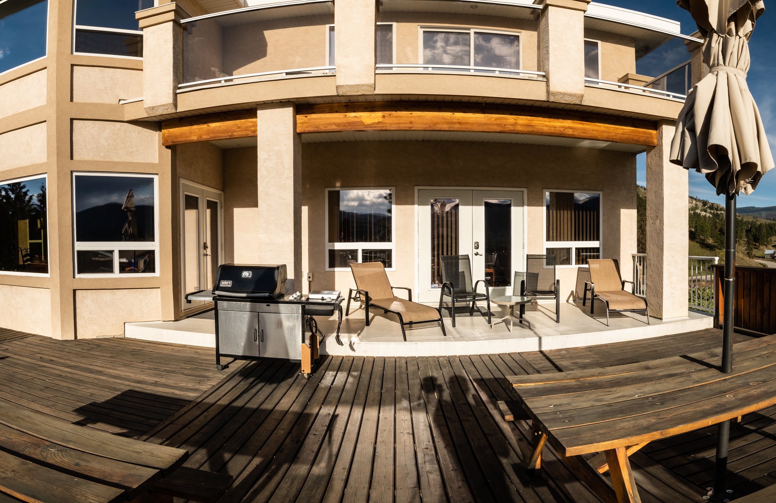 An outdoor patio on a wooden deck with lounge chairs, a small table, a barbecue grill, and a closed umbrella in front of a tan-colored house with large windows and a balcony, with mountains and trees in the background.