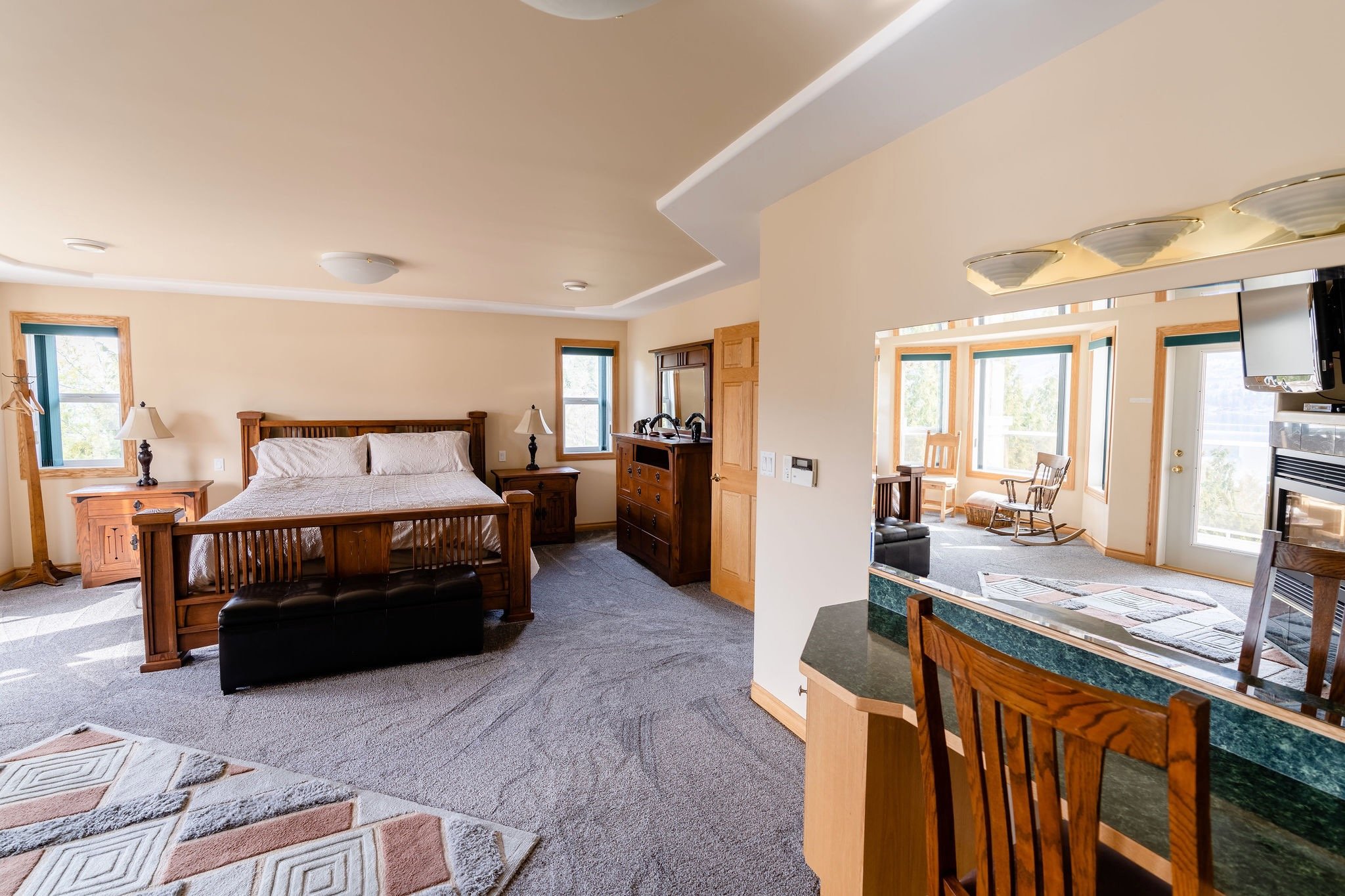 A bedroom with a wooden bed frame, two nightstands with lamps, and a dresser, with windows showing outside scenery; connected to a sitting area with rocking chairs and a glass door.