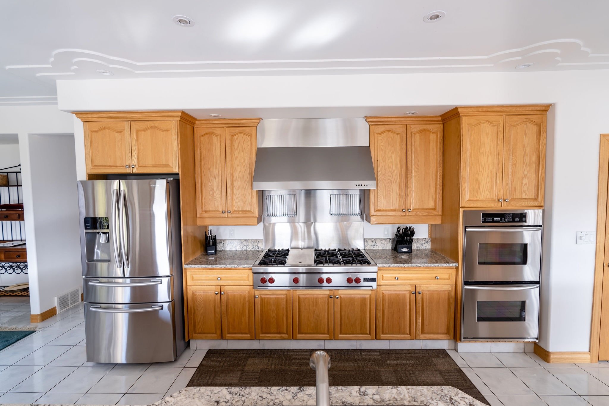 Kitchen with wooden cabinets, stainless steel refrigerator, gas stove with stainless steel range hood, and double oven, granite countertops, and tiled floor.