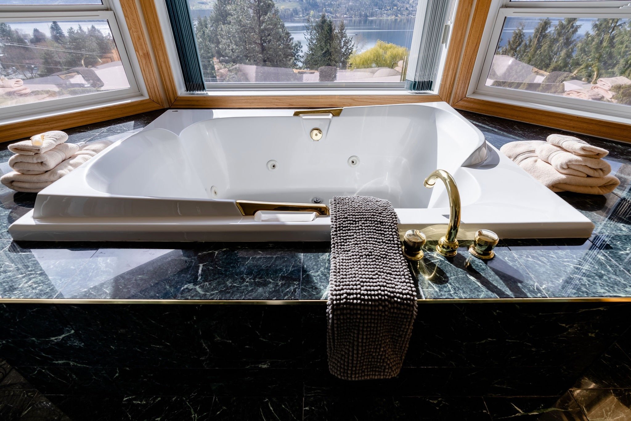 A luxurious bathroom with a corner bathtub overlooking a lake, surrounded by three windows with views of trees and water. The bathtub has gold fixtures and is decorated with folded towels on the black marble countertop.