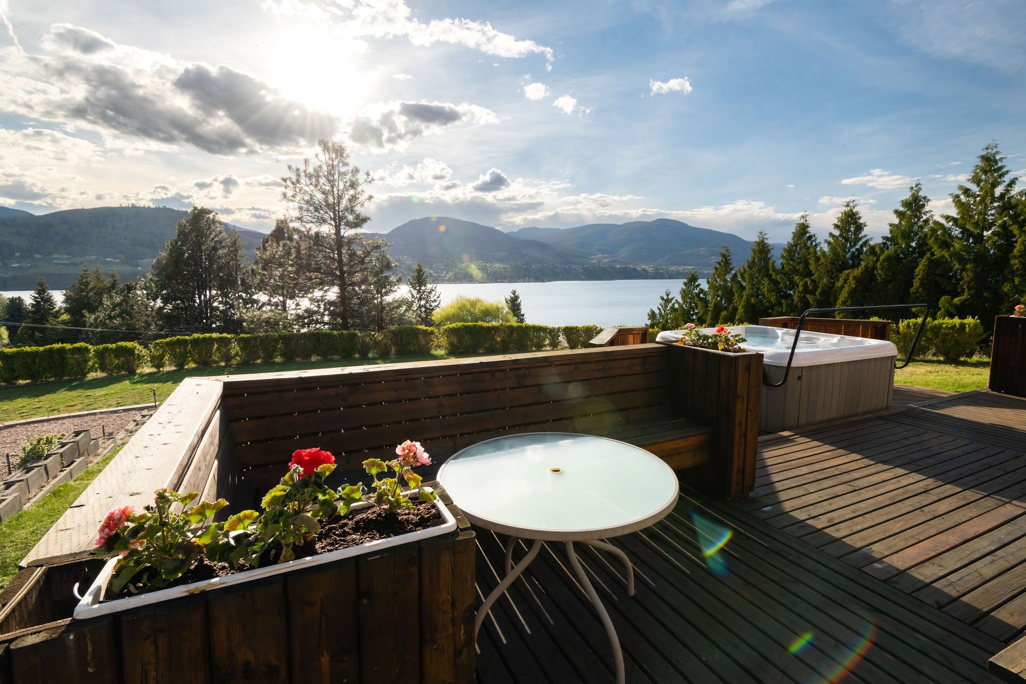 A backyard deck overlooking Skaha Lake with green hills and trees, hot tub, flower planter, and outdoor table, under a partly cloudy sky.
