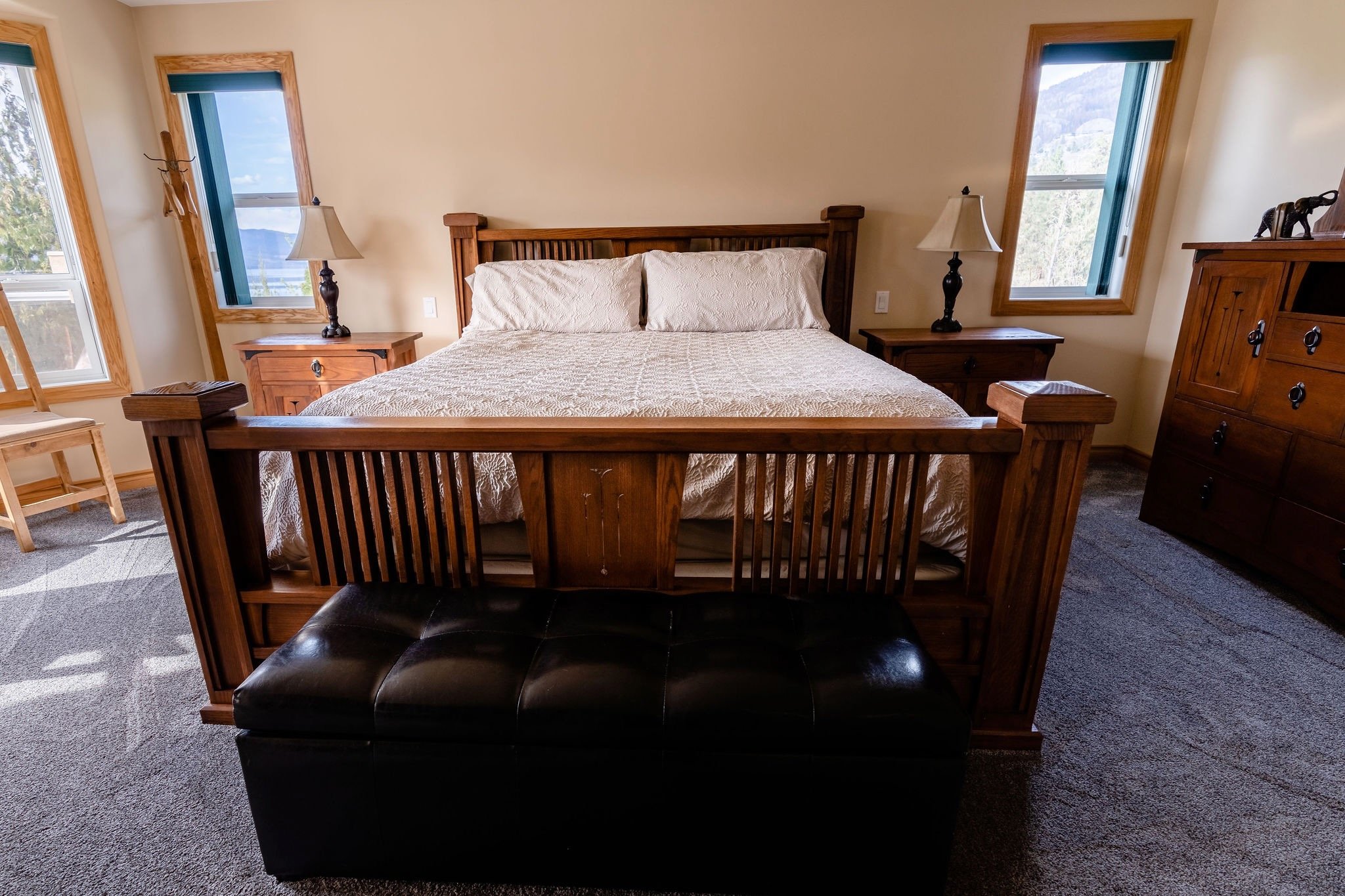A cozy bedroom with a wooden bed frame, two nightstands with lamps, two windows with blue shades, and a dark leather ottoman at the foot of the bed.