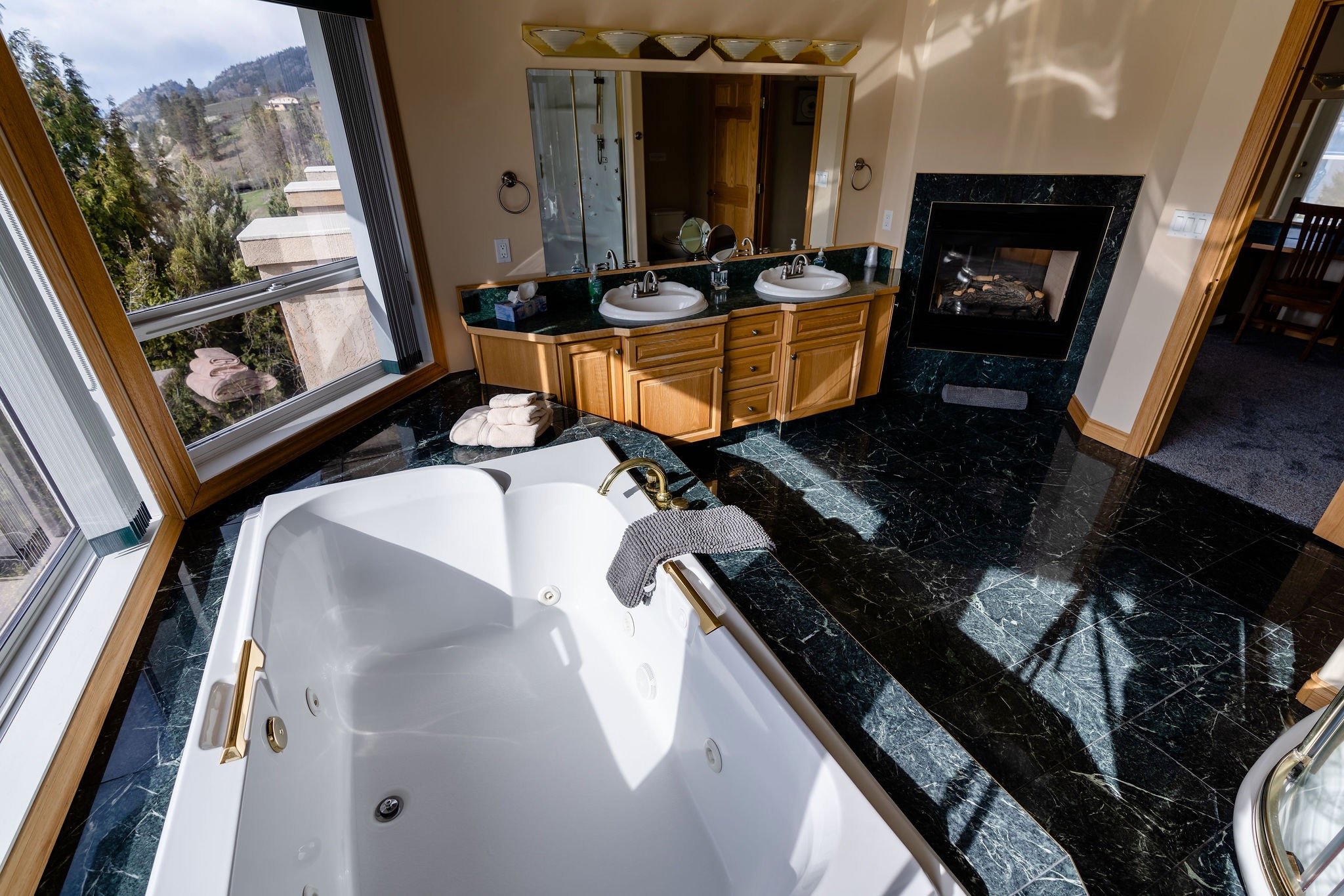 A luxurious bathroom featuring a large corner bathtub beneath two windows, a double vanity with two sinks and a mirror, a fireplace with a black marble frame, and granite countertops with wooden cabinetry.
