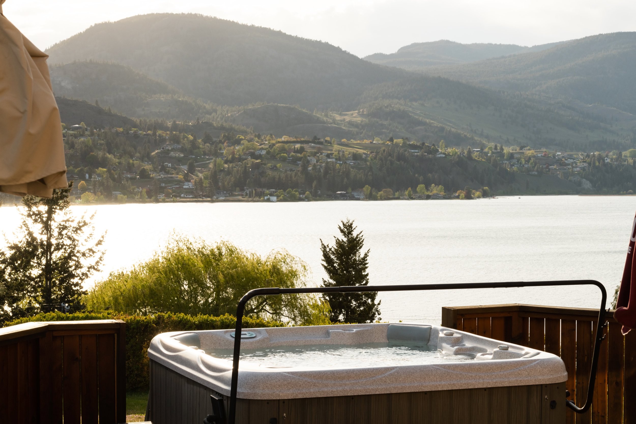 Hot tub on a wooden deck overlooking a calm Skaha Lake with green trees and hills in the background during the day.