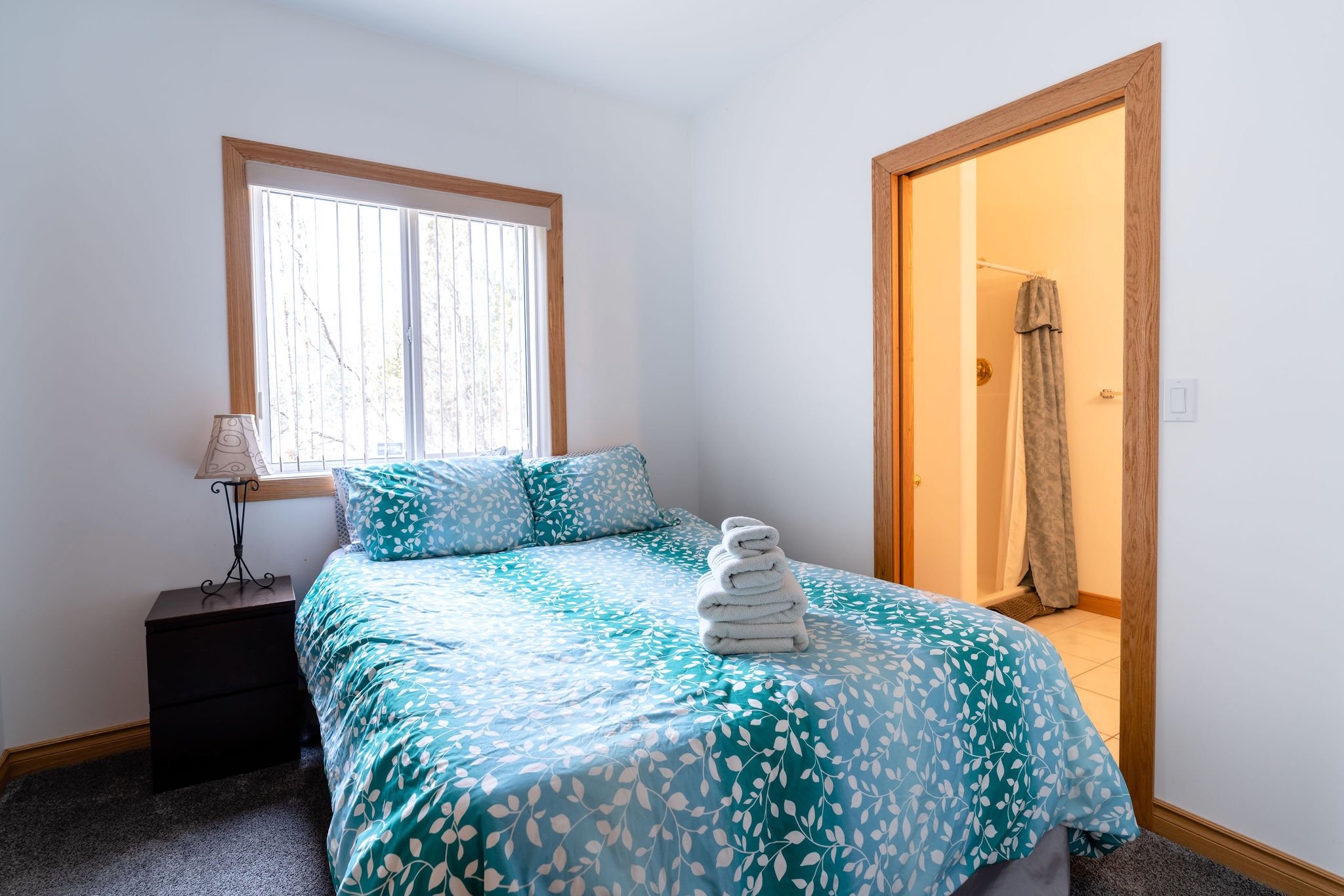 A bedroom with white walls, wooden trim, a window with blinds, a bed with a blue and white leaf-patterned bedspread and pillows, a black nightstand with a lamp, and a small closet with an open door revealing an ironing board and a curtain.