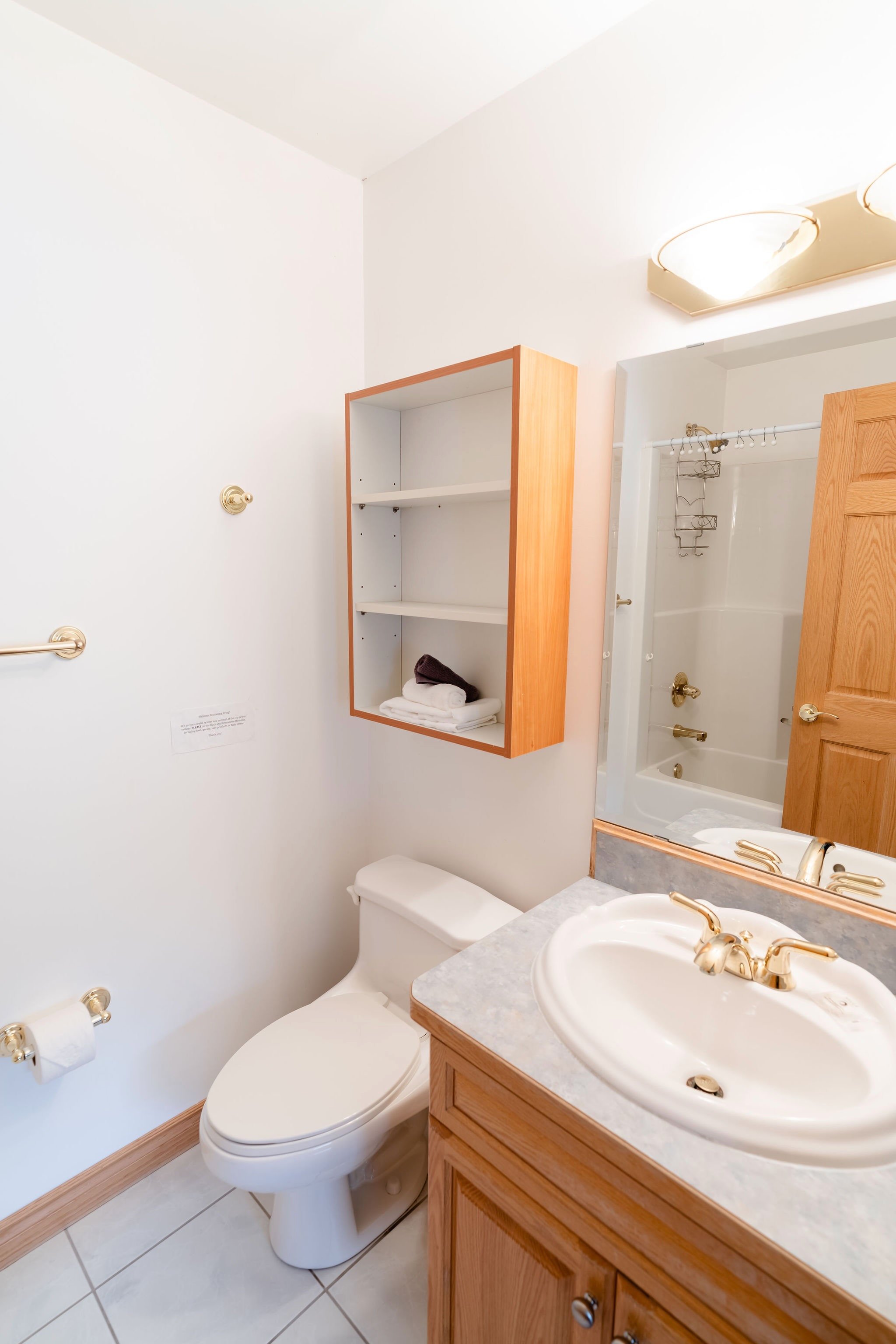 Bathroom with a toilet, wooden cabinet with open shelves, bathroom sink with gold fixtures, mirror, and a bathtub with shower curtain rod.