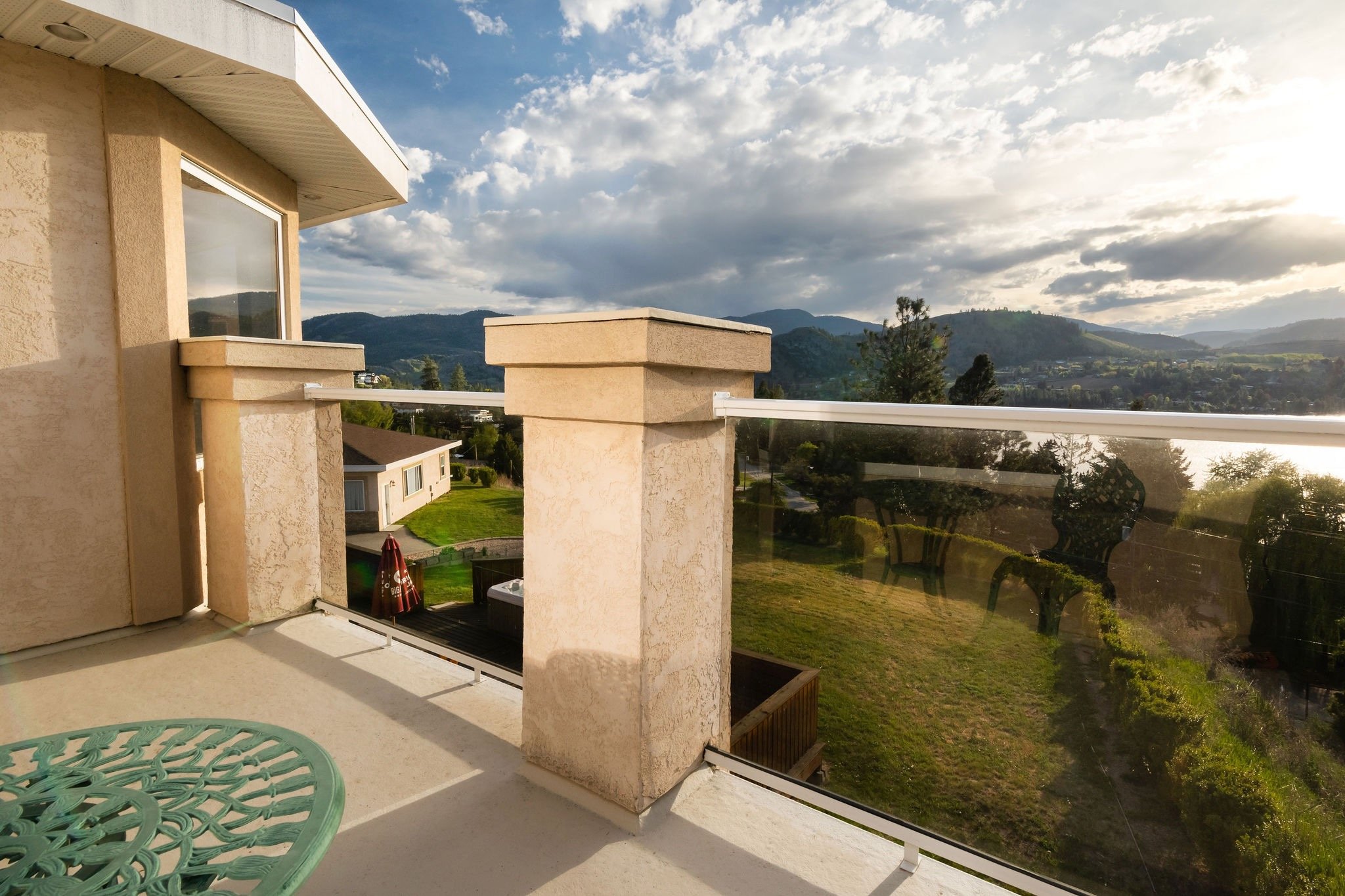 Balcony with glass railing overlooking green yard and hills in the distance, with a cloudy sky.