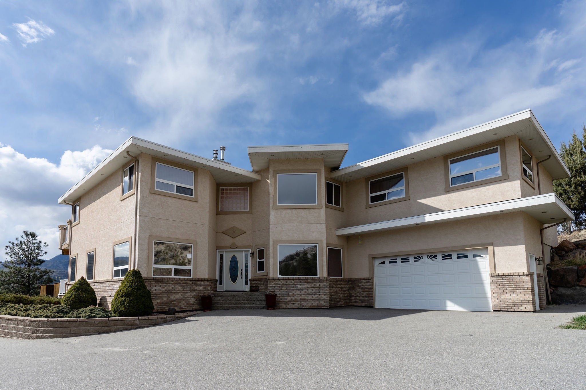 SKaha Vista Lodge front entrance exterior with beige walls, brick accents at the base, multiple large windows, a white double-garage door, a front door, and a driveway, surrounded by a landscaped yard with bushes and trees, under a partly cloudy sky.