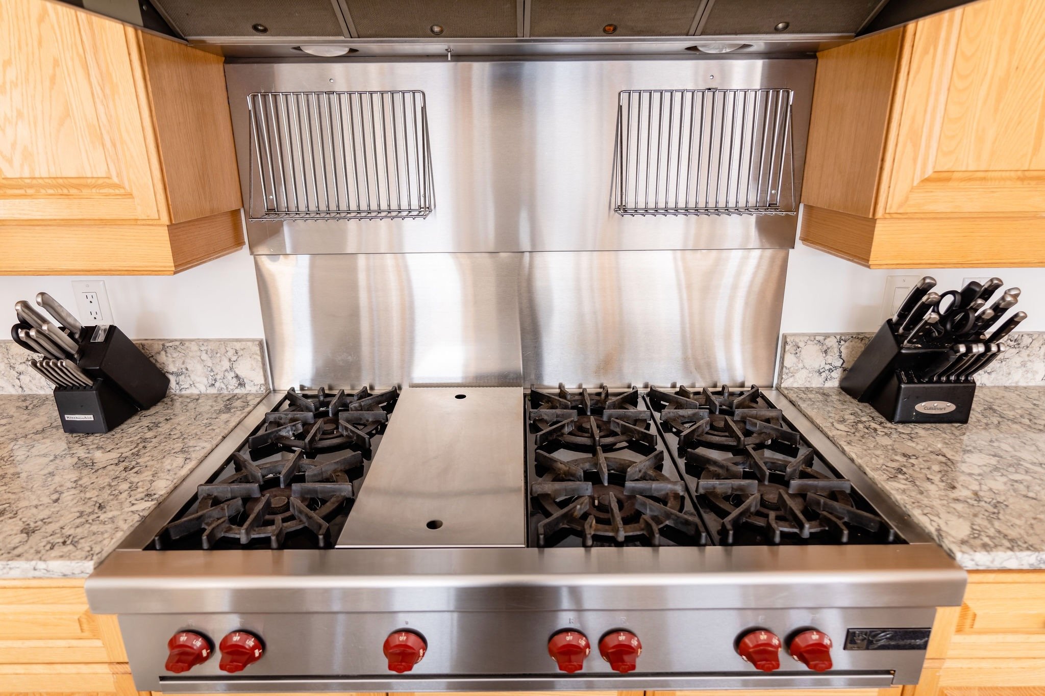 Stainless steel six-burner gas stove with black grates, red control knobs, surrounded by granite countertops and wooden cabinets, with knives in black knife blocks on either side.