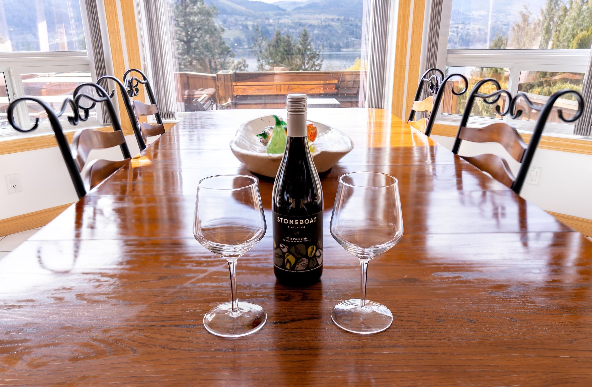 A dining room with a wooden table, two empty wine glasses, and a bottle of Stoneboat Pinot Noir in the center. There is a decorative bowl in the background, and large windows revealing a scenic view of trees, a lake, and distant mountains.