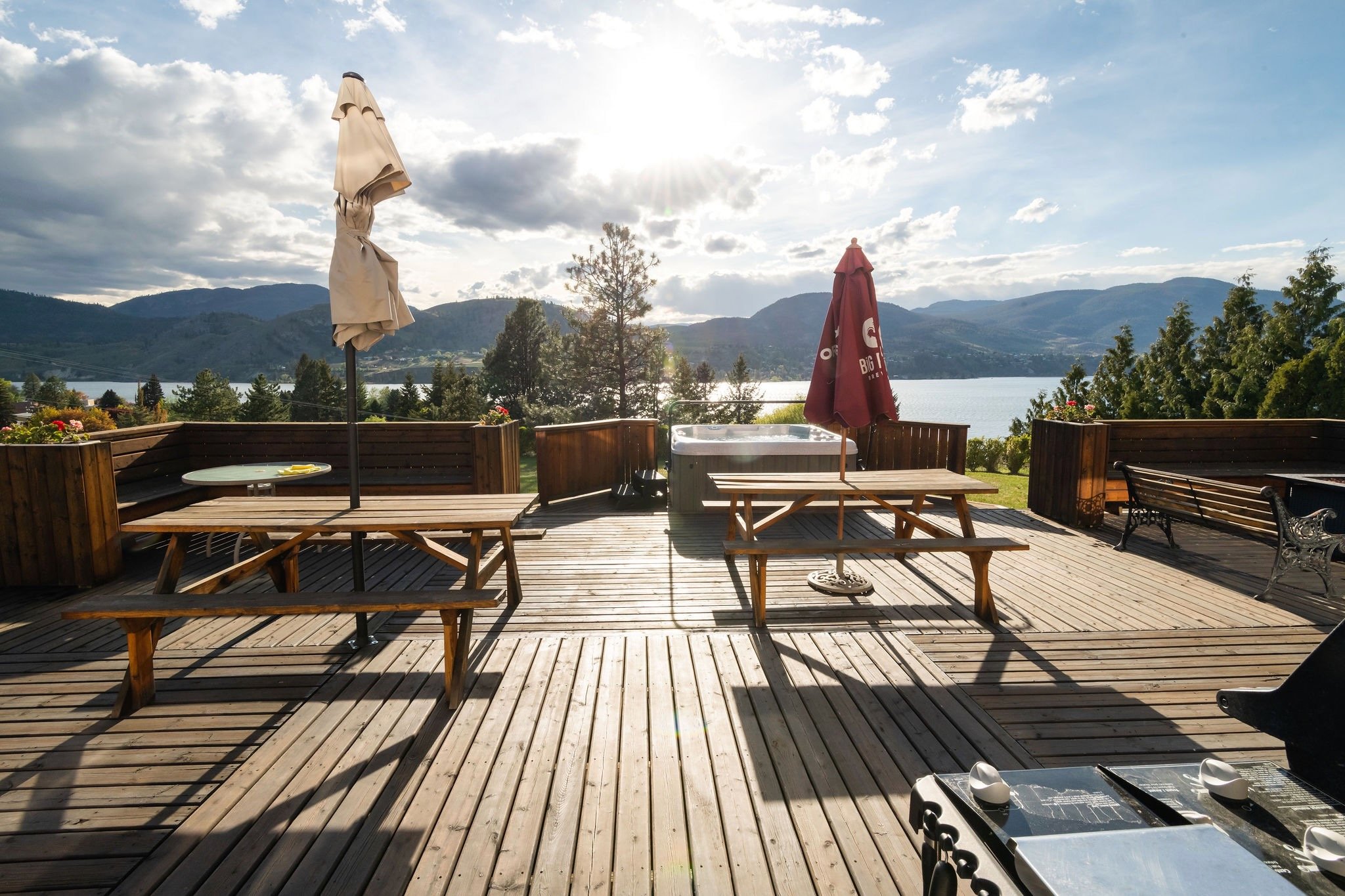 Wooden deck overlooking Skaha Lake with mountains, with umbrellas, benches, a hot tub, and a barbecue grill.