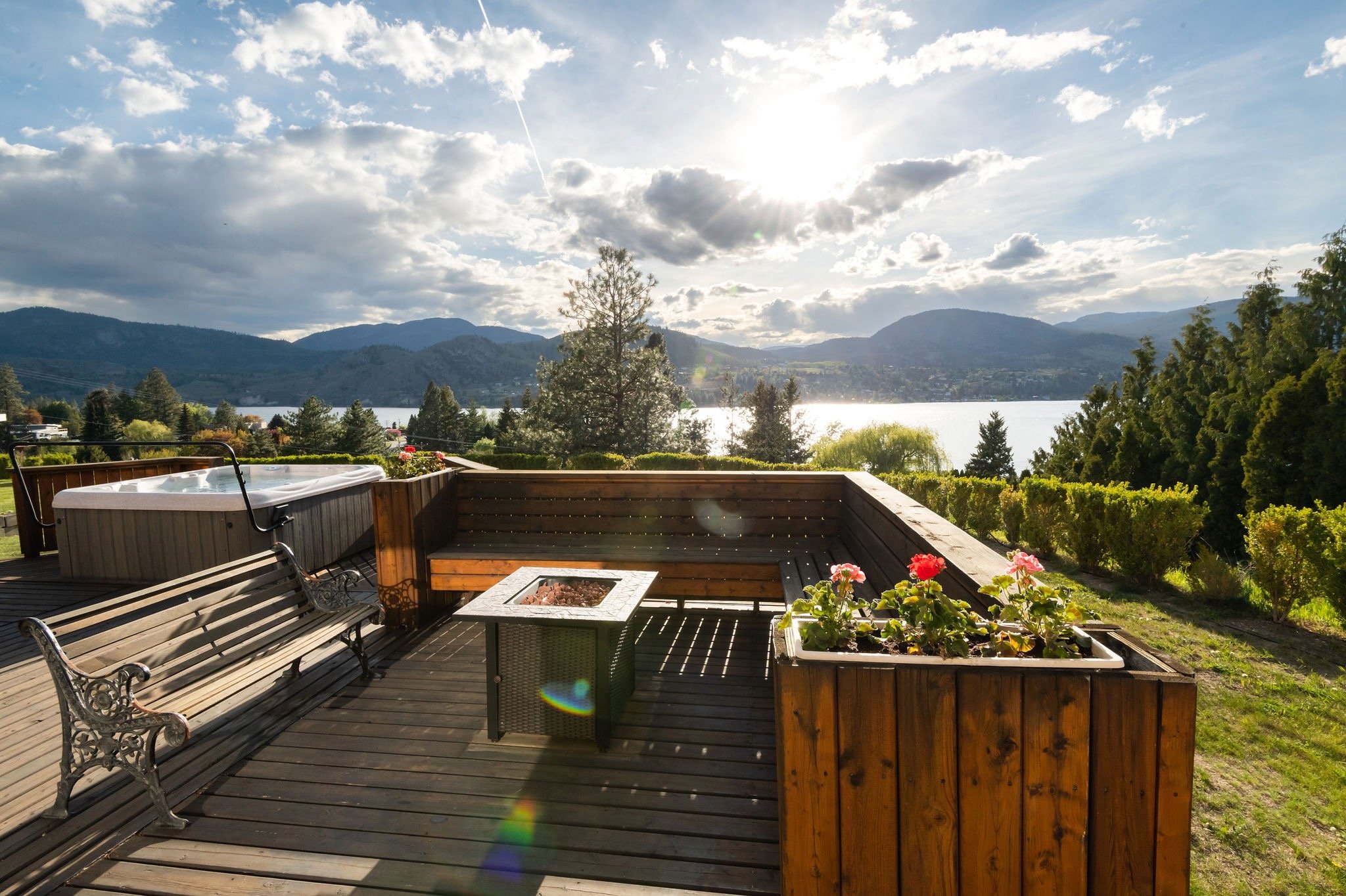 A wooden deck overlooking Skaha Lake and mountains with a hot tub, a bench, a fire pit, and flower planters under a partly cloudy sky.