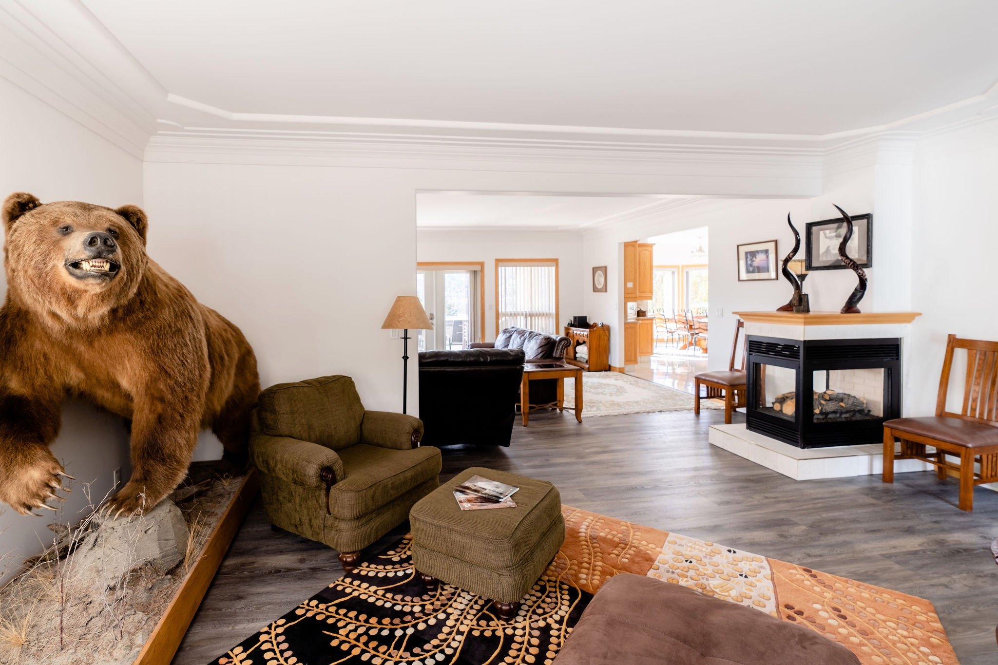 Living room with a taxidermy kodiak brown bear mounted on the wall, a brown armchair with an ottoman, a fireplace with decorative objects and framed pictures above it, and other seating and furniture in the background.