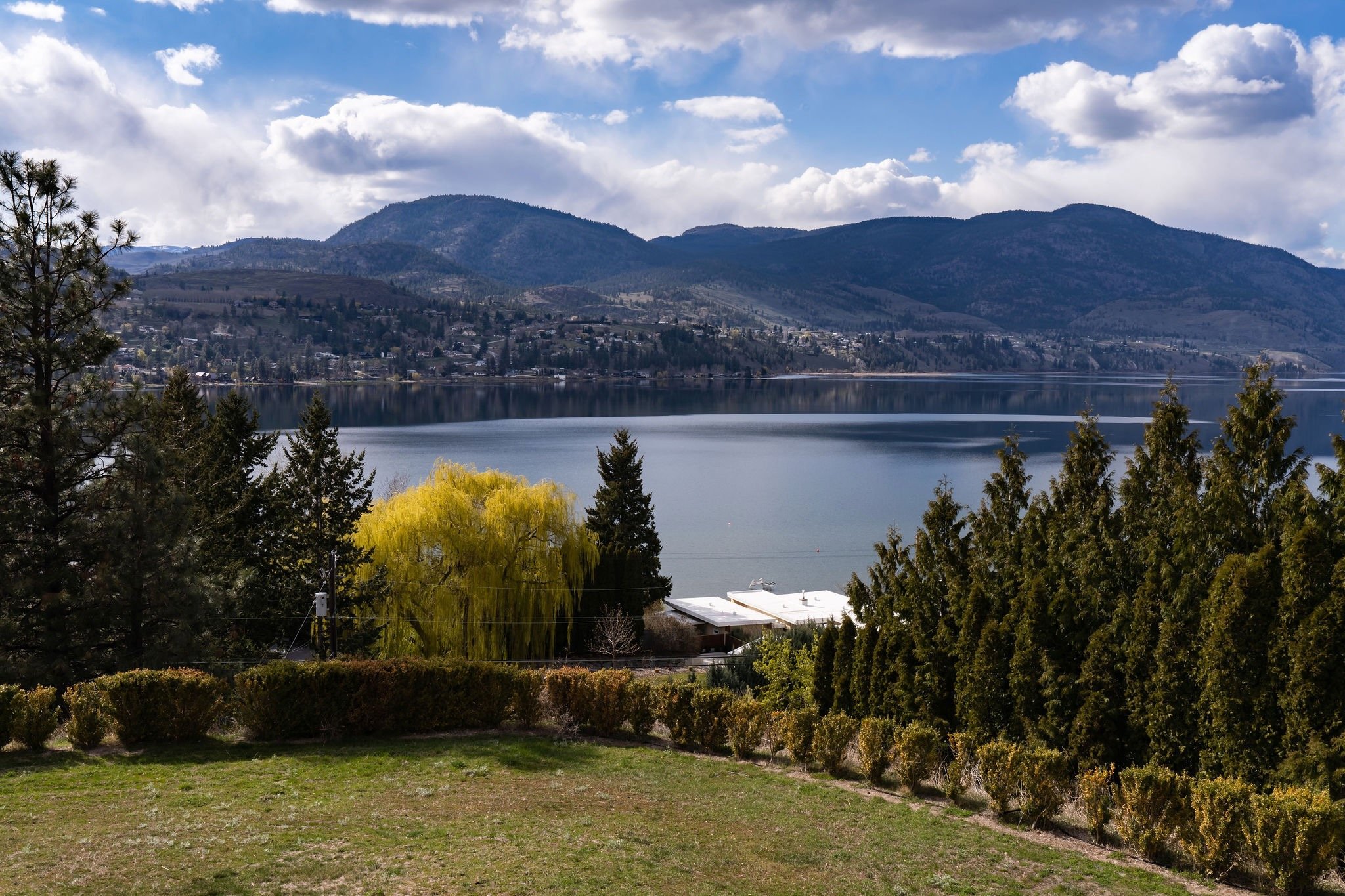 Scenic view of a Skaha Lake surrounded by trees and mountains under a partly cloudy sky.