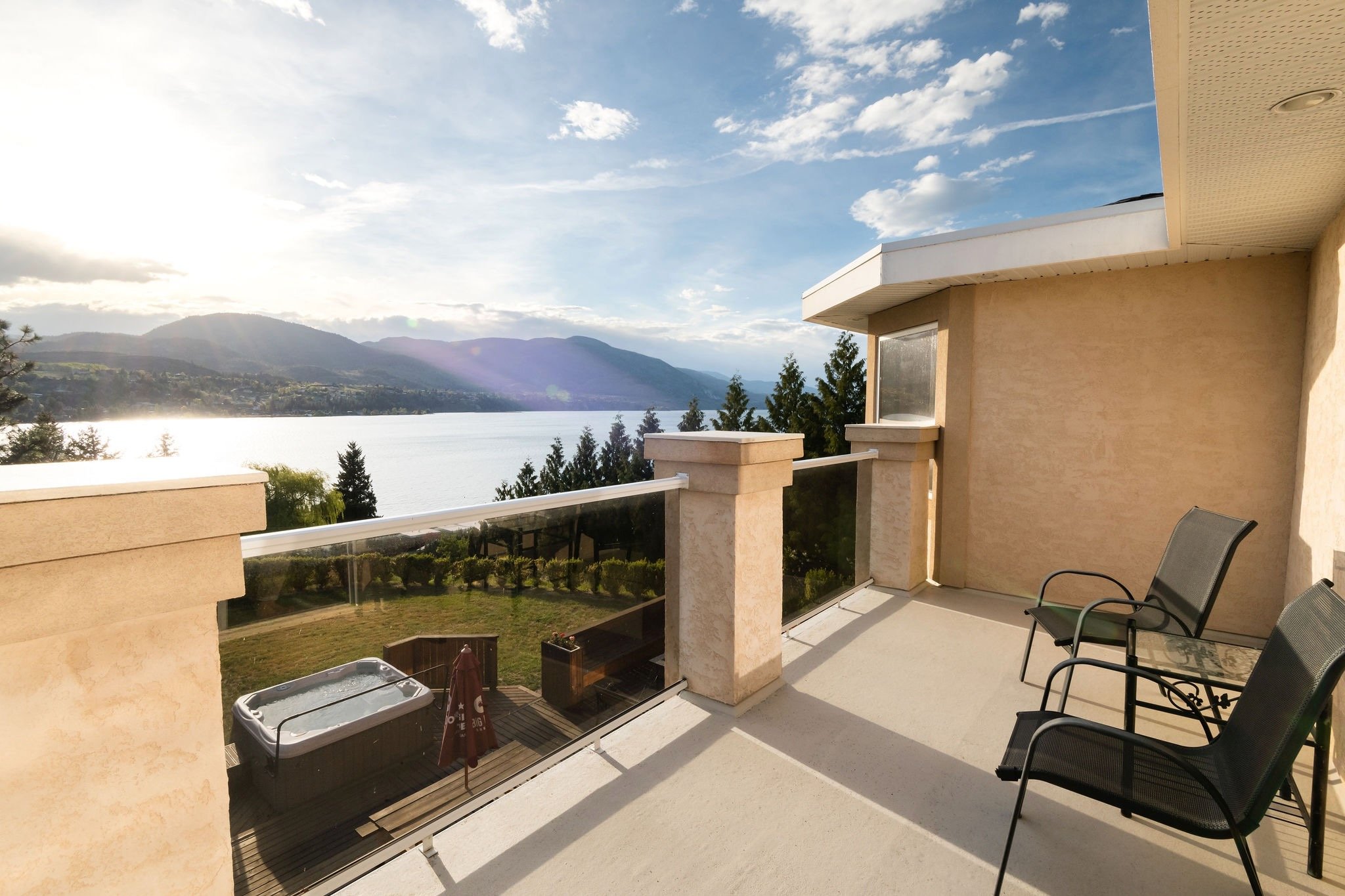 Balcony overlooking Skaha Lake and mountains with a hot tub, two chairs, and a view of a partly cloudy sky.