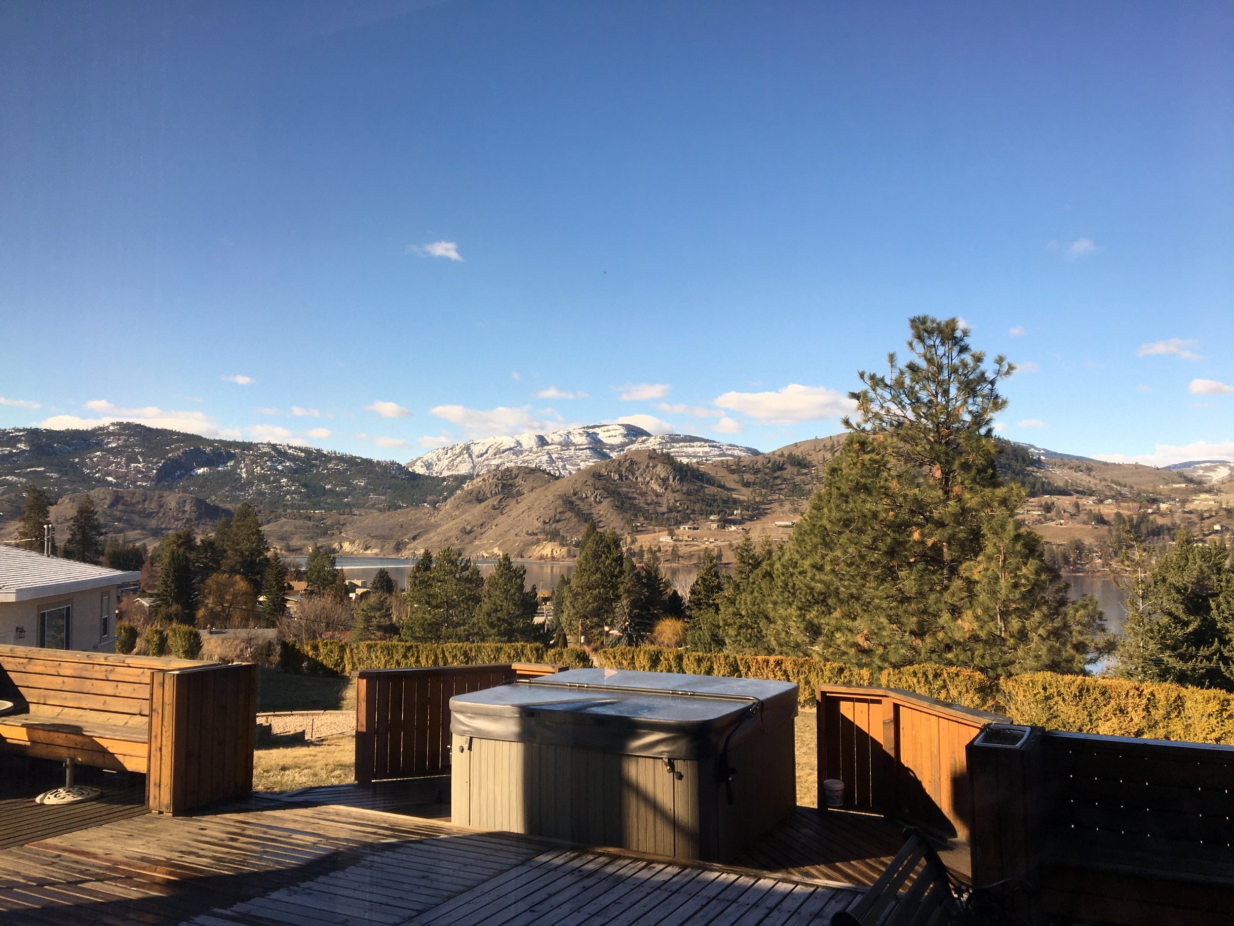 Sunny backyard with wooden deck, hot tub, trees, distant mountains with snow, and a lake under a blue sky with scattered clouds.