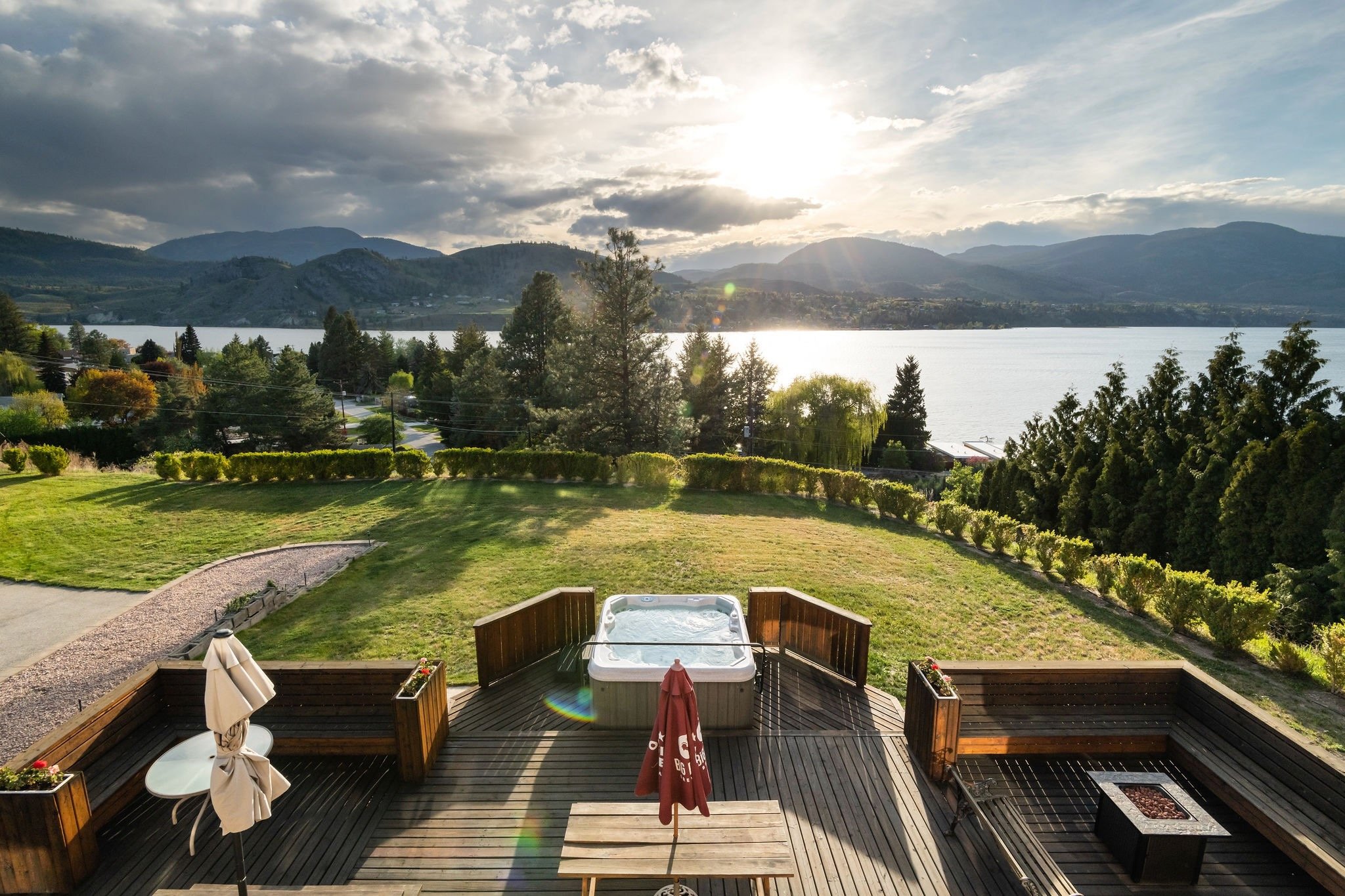 A backyard deck overlooking Skaha Lake with mountains in the background. The deck has a hot tub, a table with an umbrella, and fire pit.