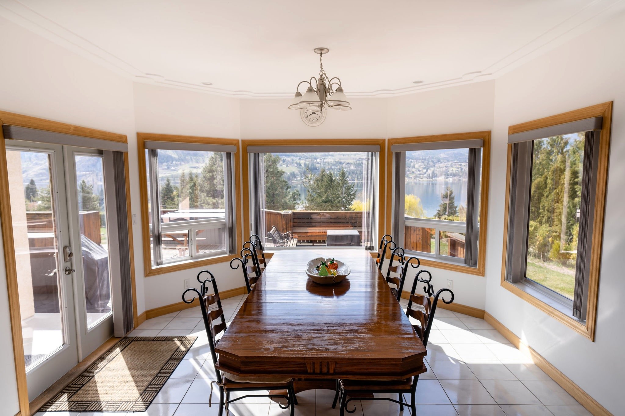 Dining room with a large wooden table and eight black chairs, surrounded by windows showing a lake and trees outside, with a chandelier hanging from the ceiling.
