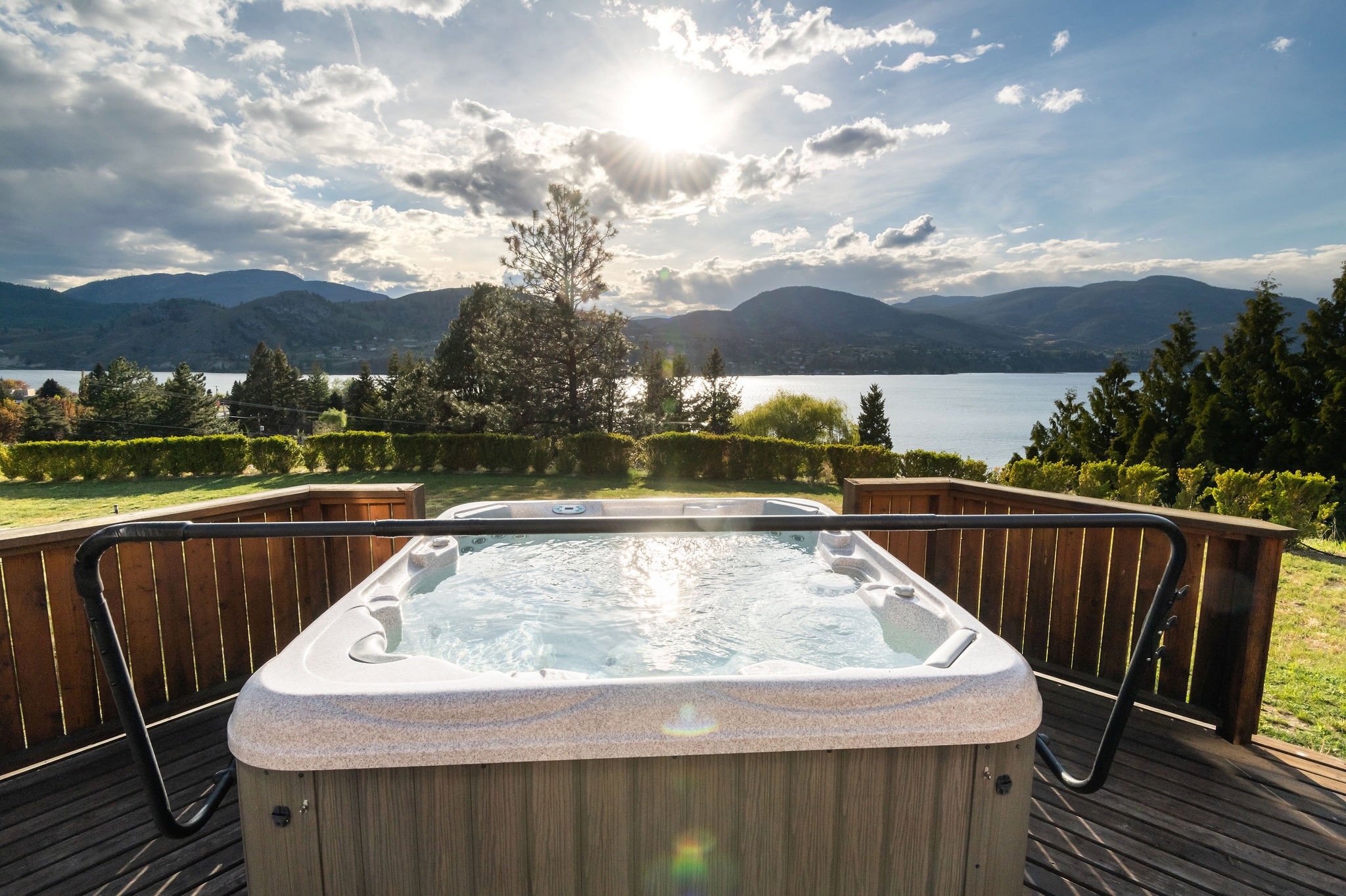 Hot tub on a wooden deck overlooking Skaha Lake with mountains in the background, under a partly cloudy sky with the sun shining