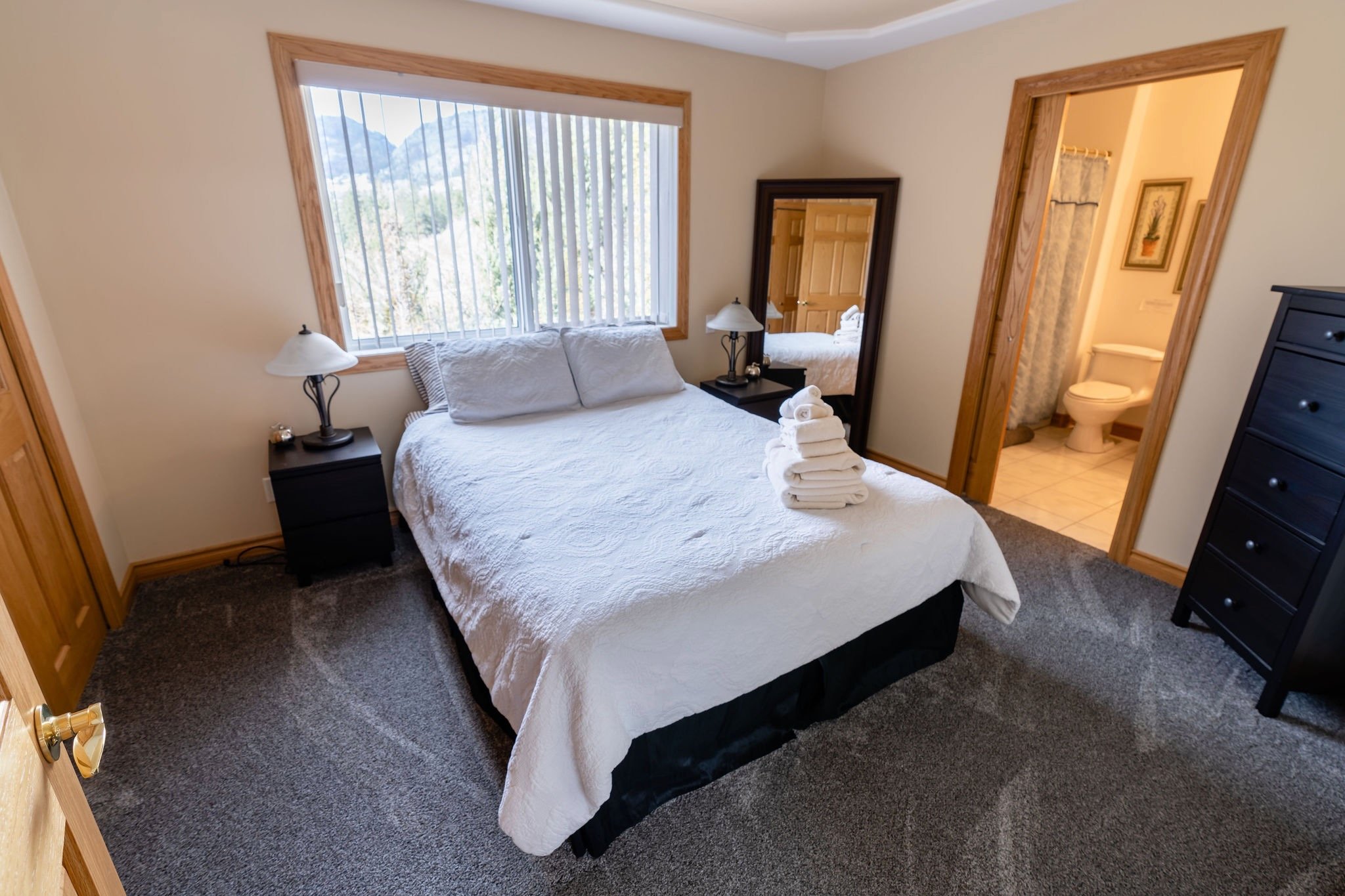 Room with a bed, nightstands, lamps, a window with vertical blinds, a mirror, and a bathroom with a toilet in the background