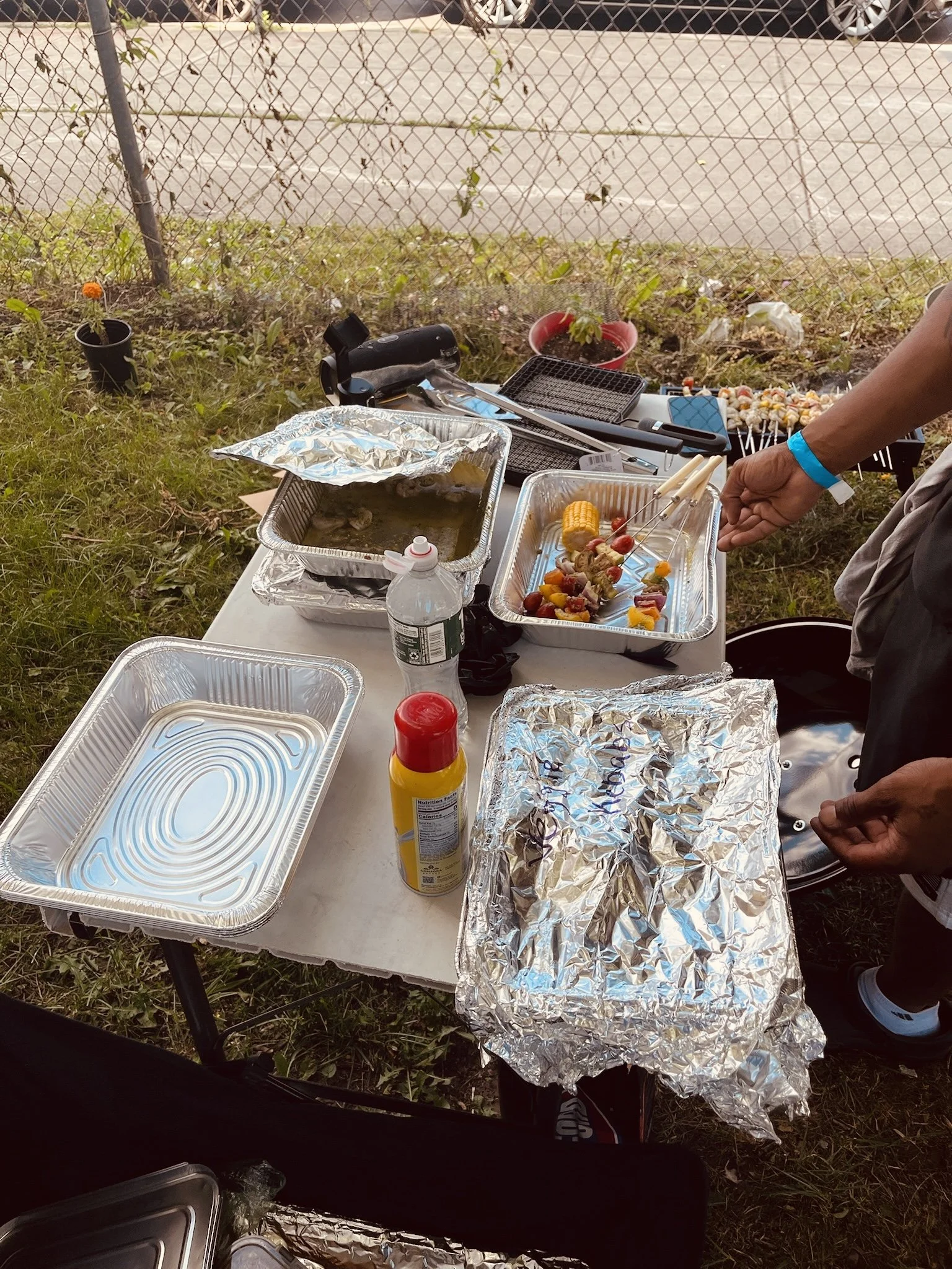 Outdoor barbecue setup with aluminum trays of food, skewers, and grilling tools on a table.