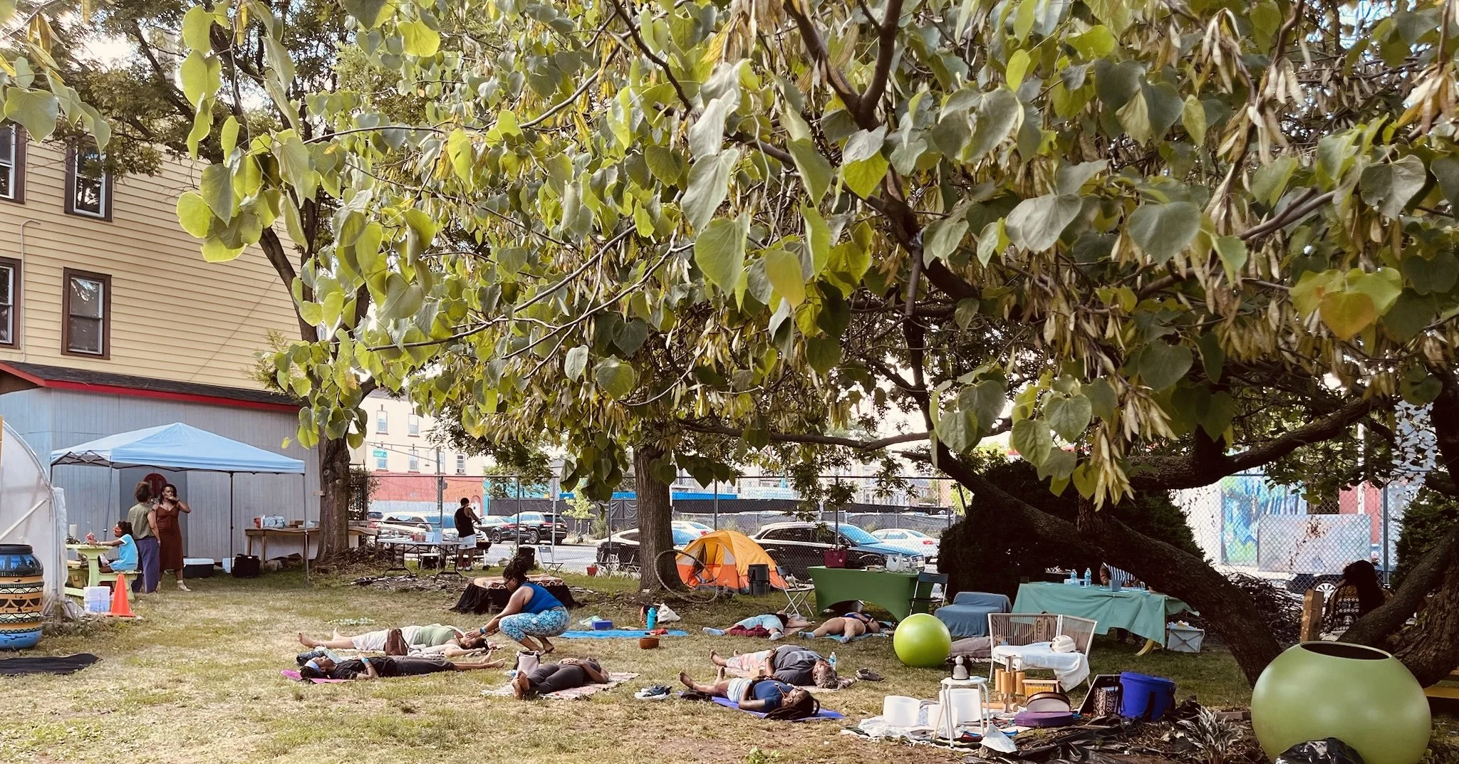 People practicing yoga outdoors under a large tree in a park, with some sitting and some lying down on yoga mats. There are tents, chairs, and coolers around, and a cityscape with buildings and parked cars in the background.