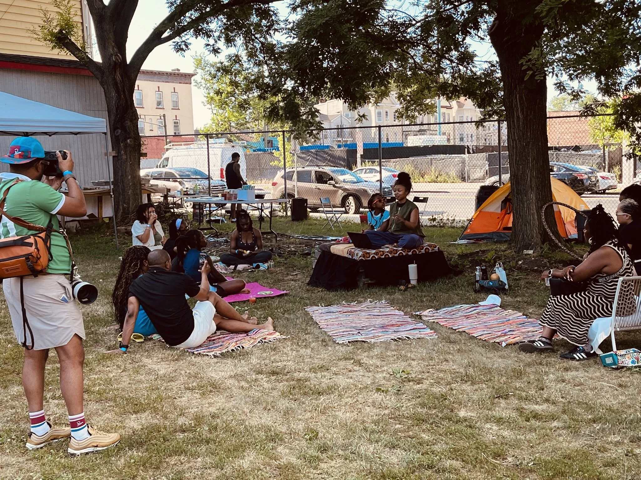 A group of people gathered outdoors under trees, sitting on colorful mats, with some standing. There are tents, a chain-link fence, parked cars, and buildings in the background. One person is taking photos, and others appear to be socializing or participating in an event.
