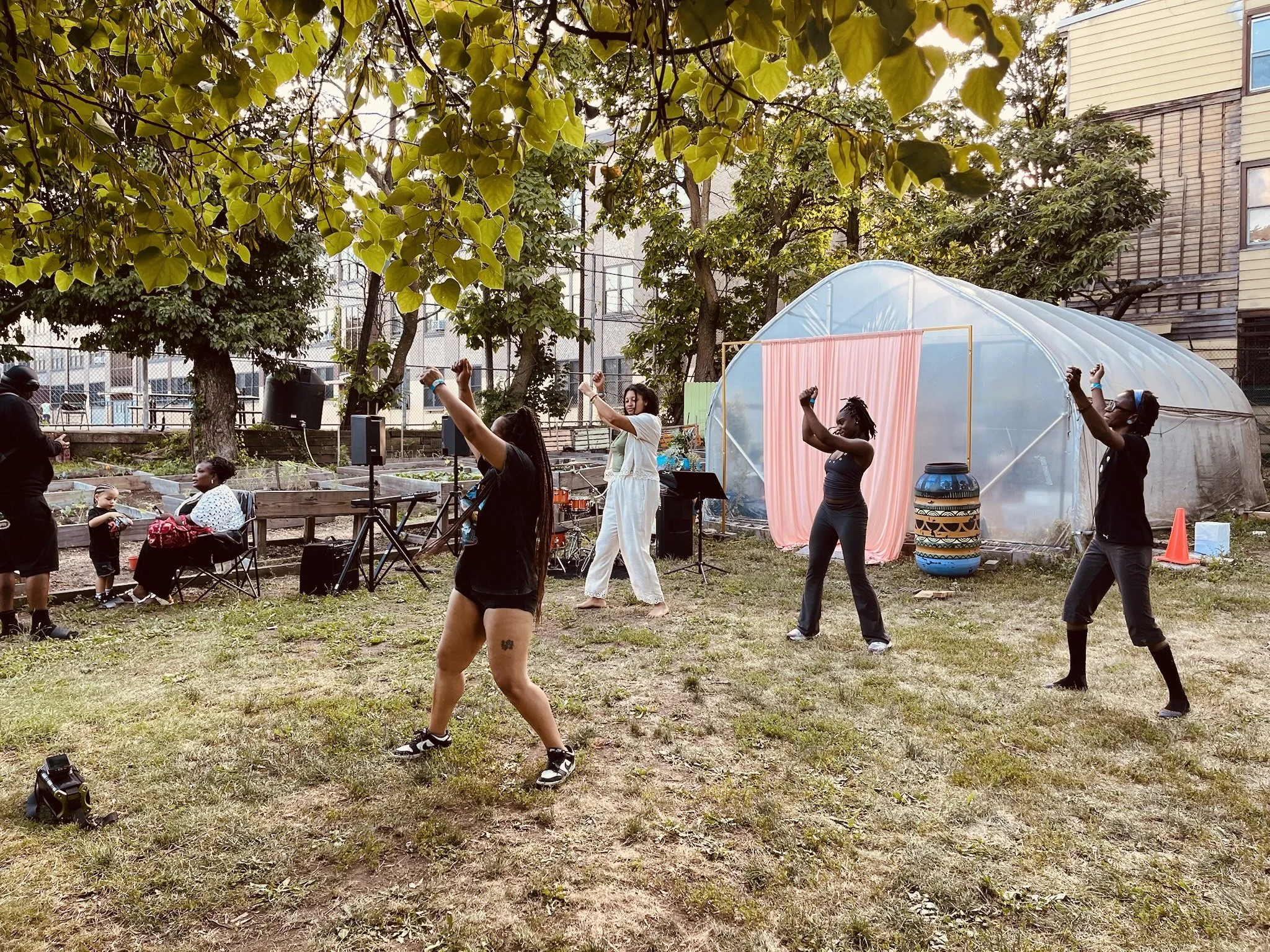 A group of people dancing on a grassy area in a park, with trees, a greenhouse, and residential buildings in the background.